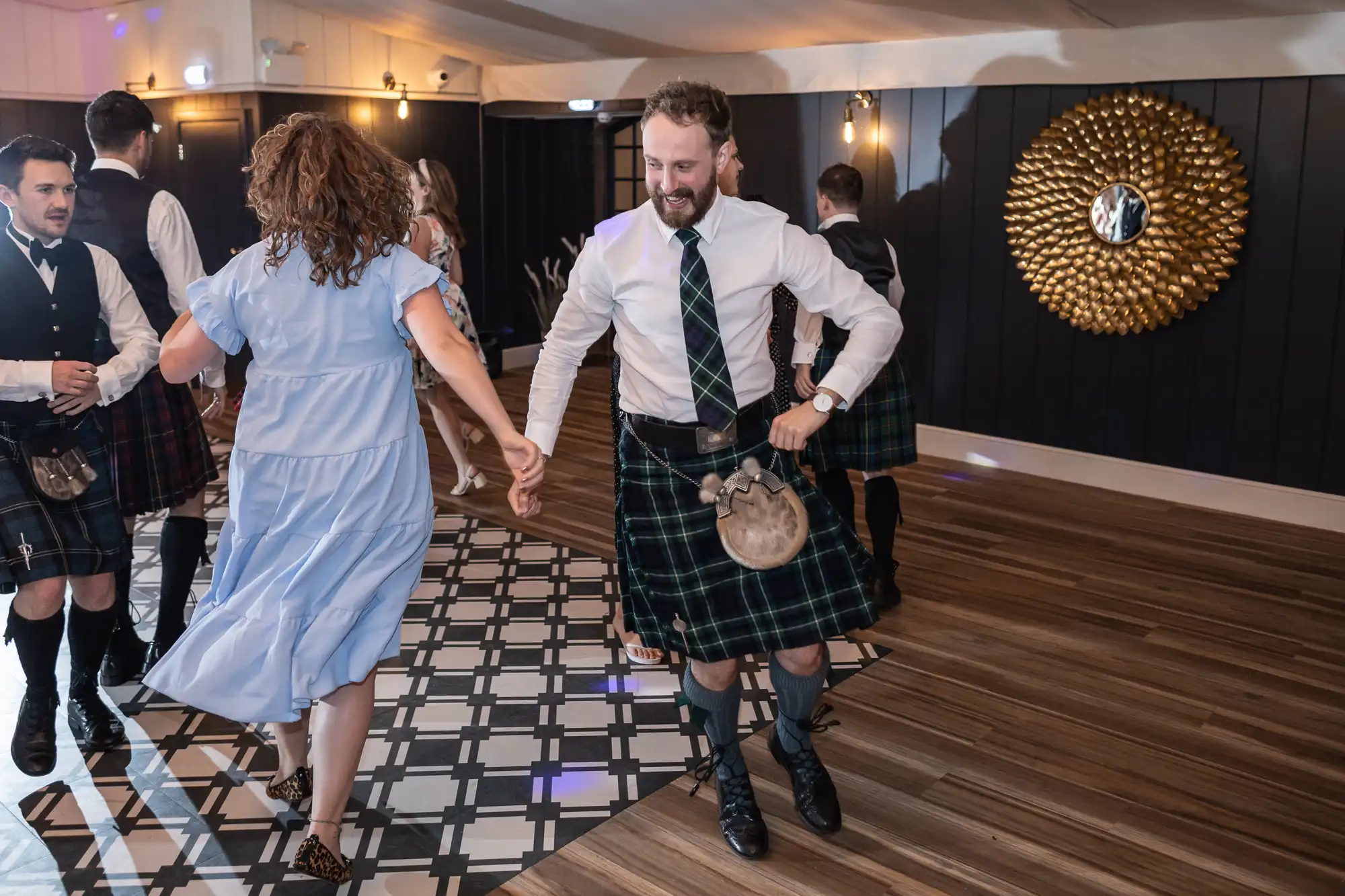 People dancing in a room with wooden floors and dark paneled walls; one man in a kilt is holding hands with a woman in a light blue dress.