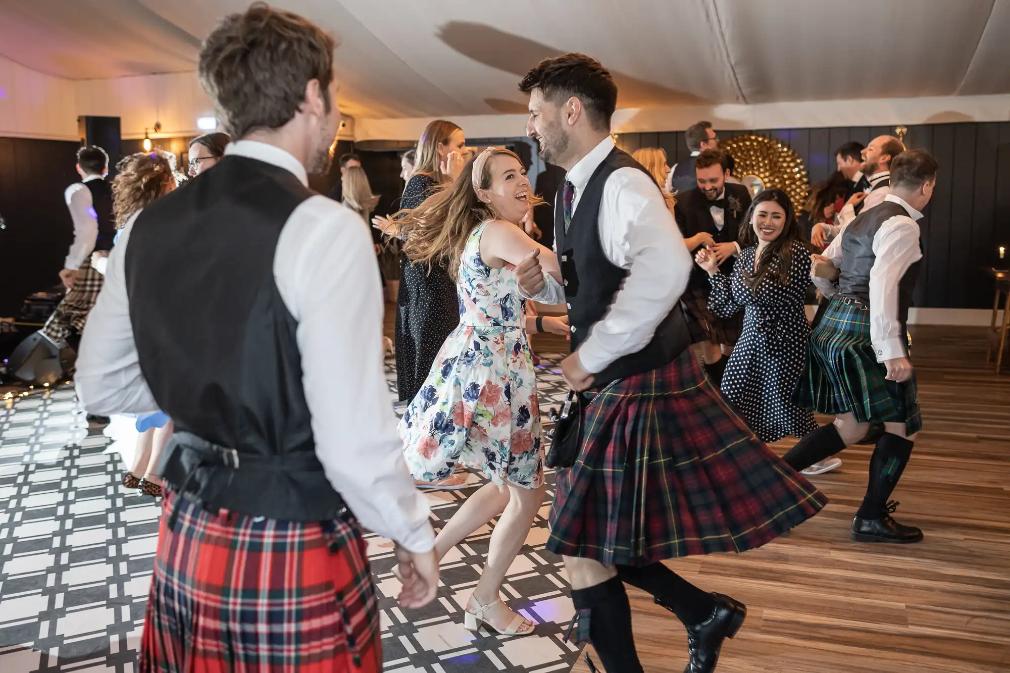 People in traditional Scottish attire dance together inside a hall, wearing kilts and vests. Some women wear dresses and skirts, and the atmosphere appears joyful and lively.