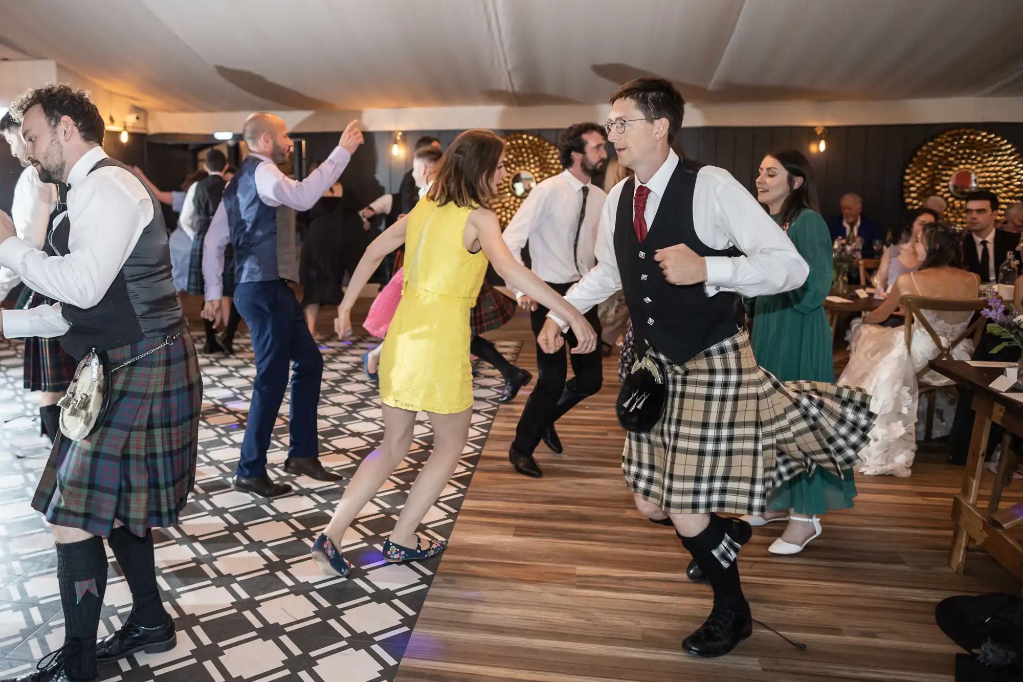 Guests in kilts and various outfits dance at a gathering with a checkered and wooden floor in an indoor venue. Some attendees are seated and observing in the background.