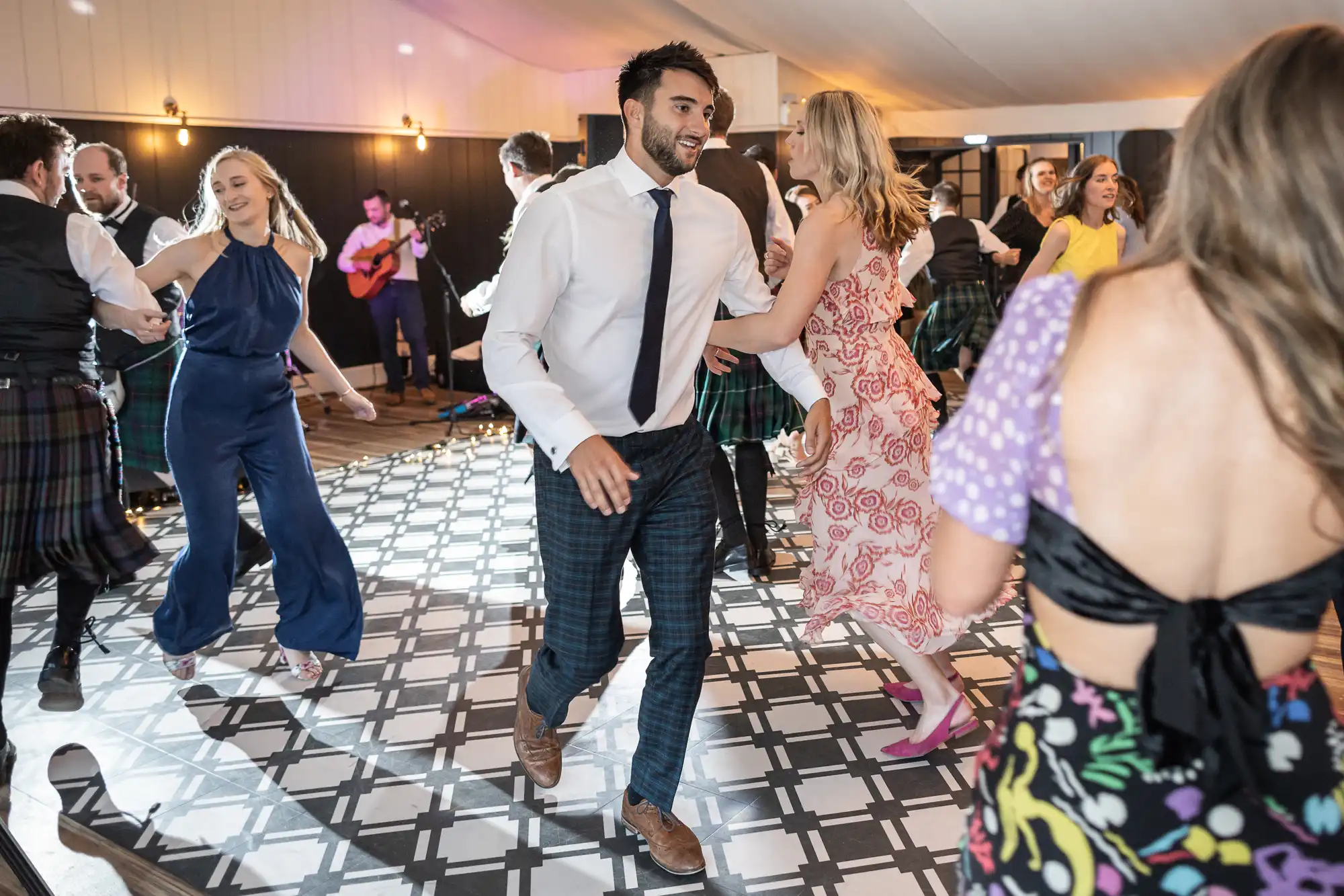 People dancing energetically at a social event, featuring a man in a white shirt and tie, and others in vibrant attire, on a patterned floor with live musicians in the background.