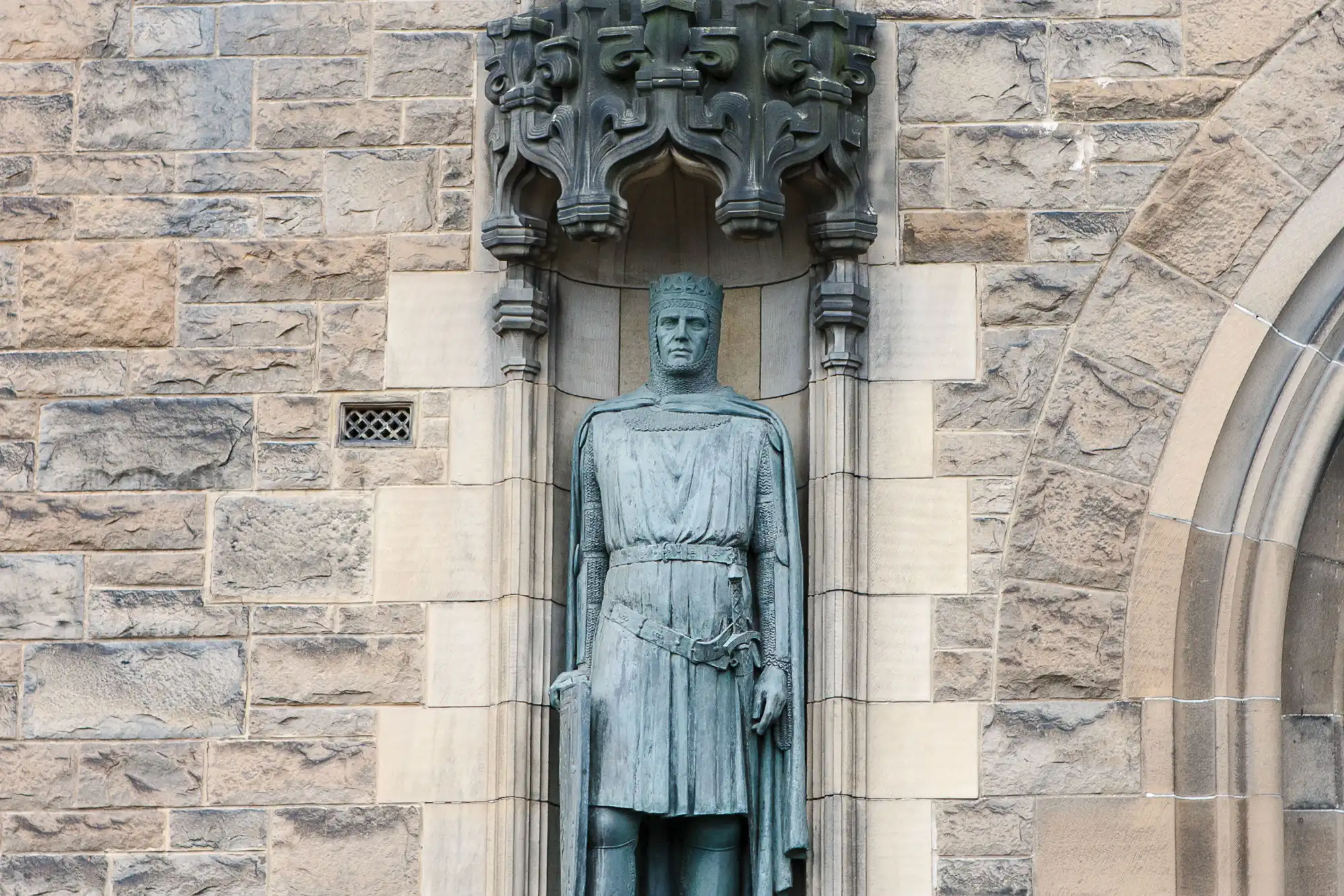 Stone statue of a medieval knight in full armor, set into a niche with ornate gothic architectural details on a stone wall.