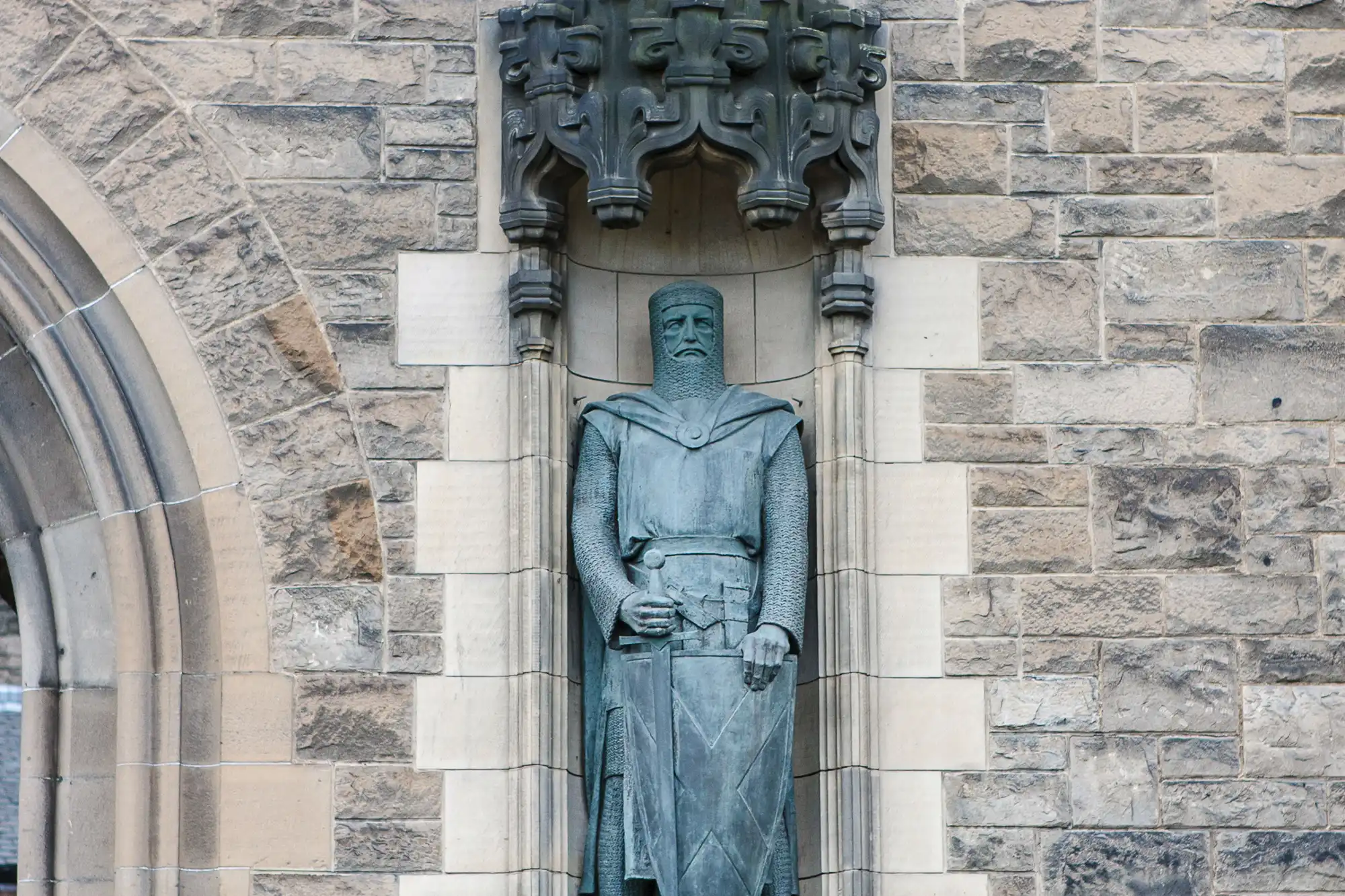 Stone statue of a medieval knight in armor, standing solemnly within a niche on a building's stone facade.