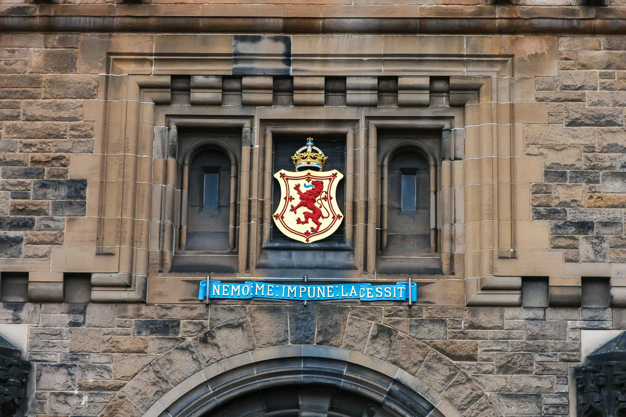 Stone facade of a building with a crest featuring a red lion and a banner reading "nemo me impune lacessit" above an arched doorway.