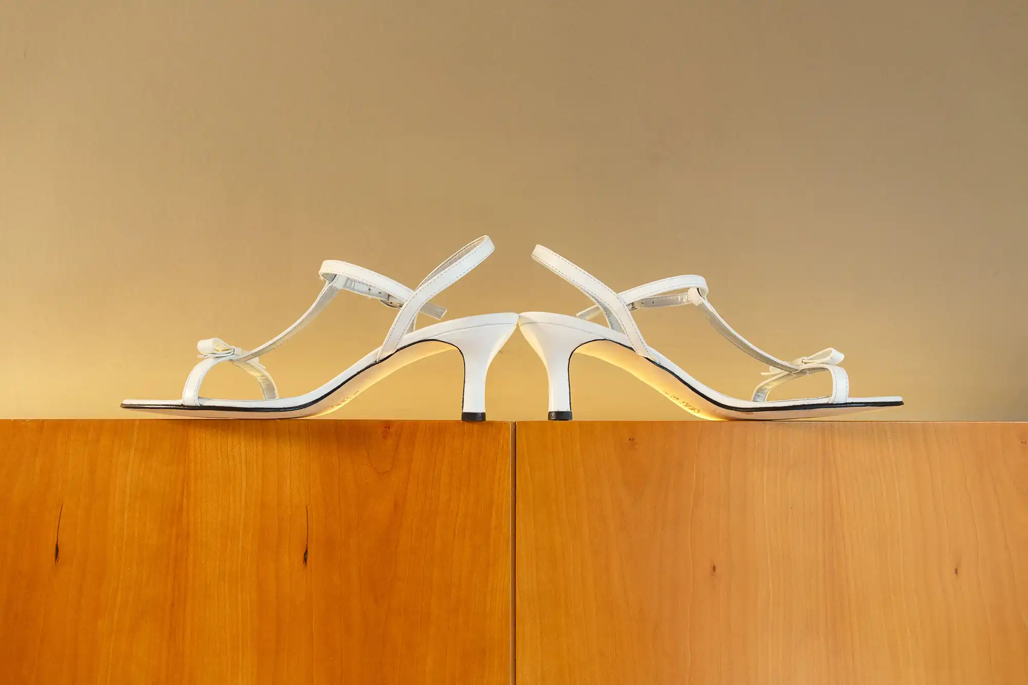 A pair of white heeled sandals with thin straps displayed on a wooden surface against an ochre background.
