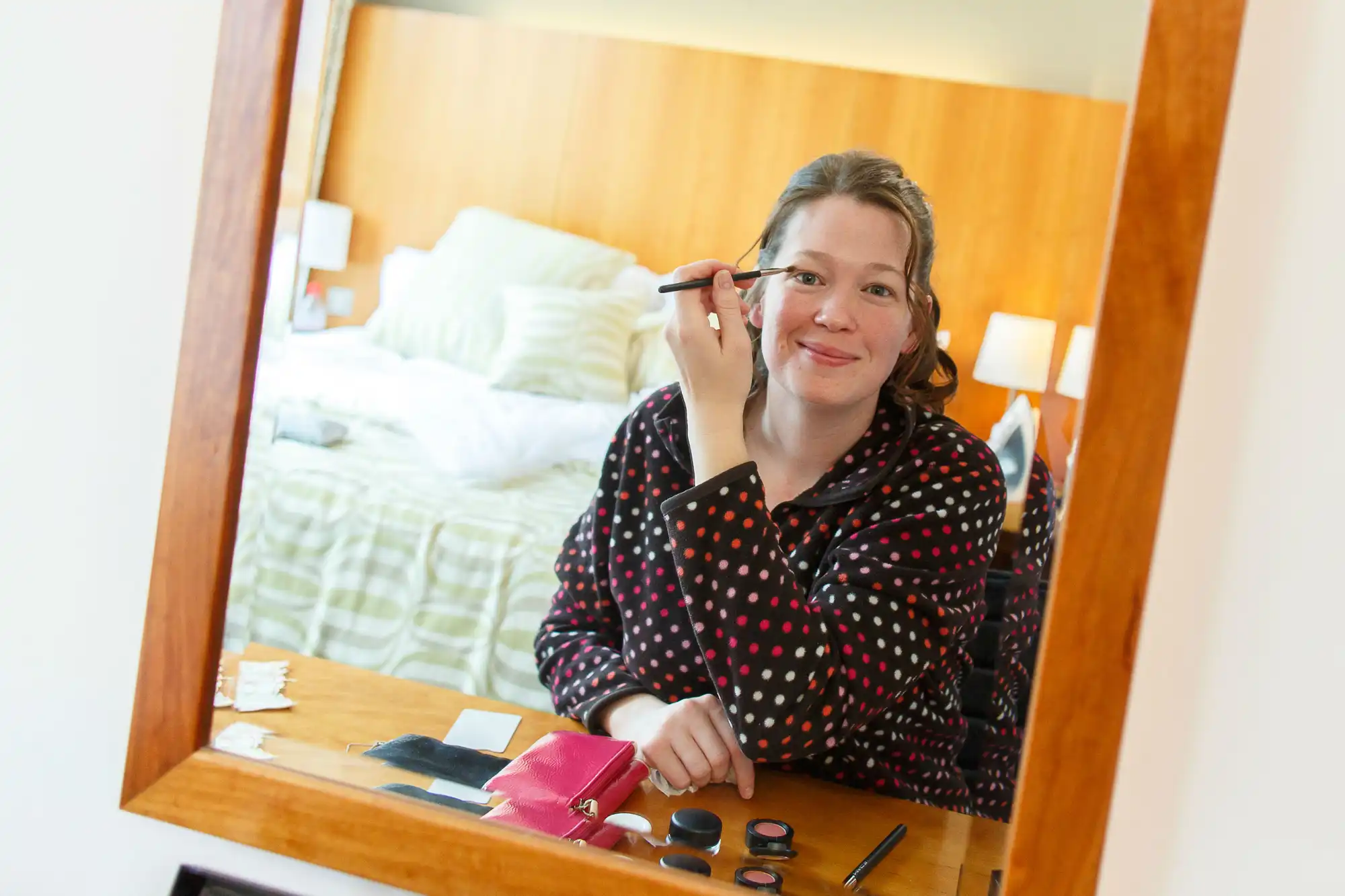 A woman applying makeup in front of a mirror, with a bed visible in the reflection.