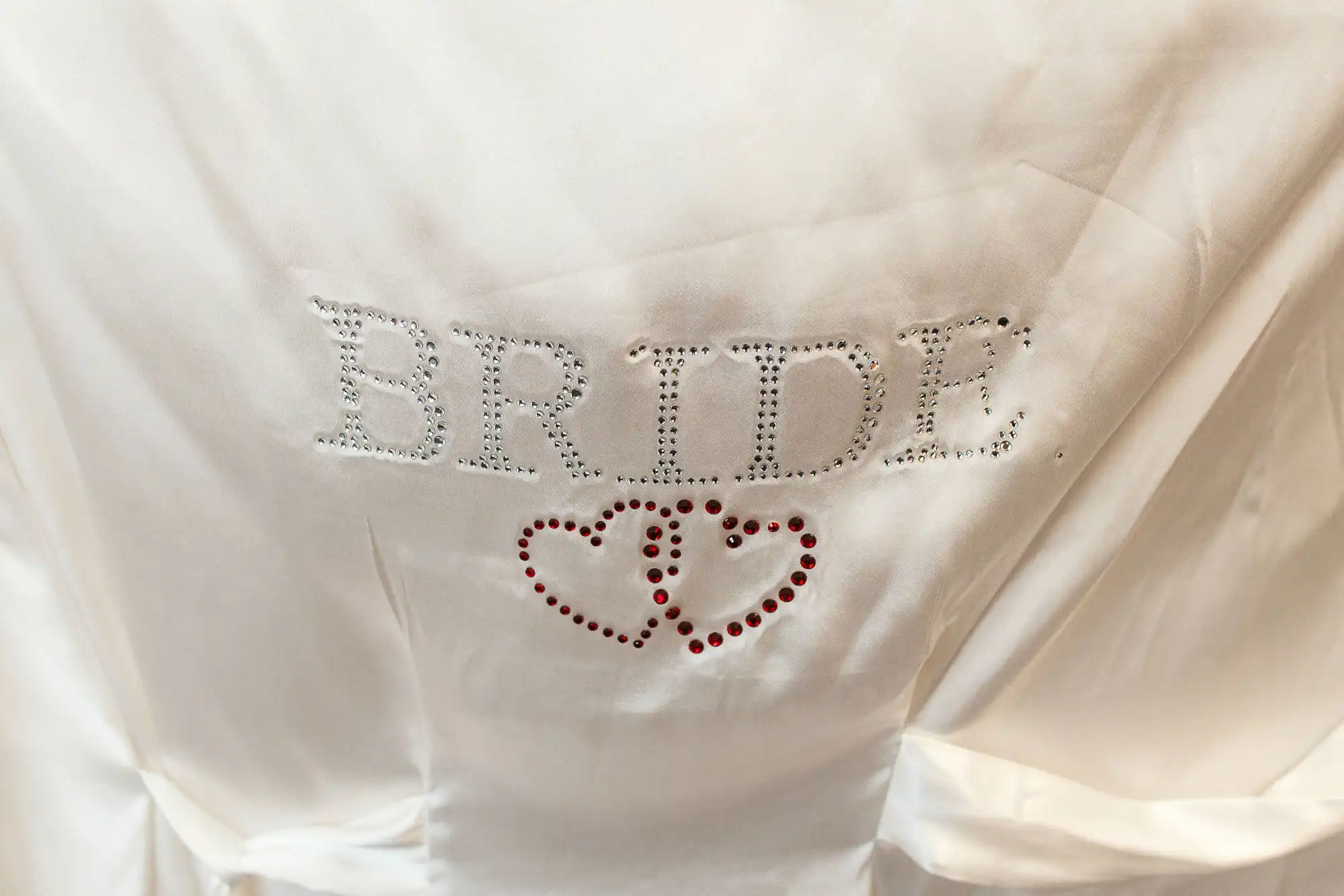 White satin robe with "bride" in rhinestones and two red hearts below, draped and close-up.