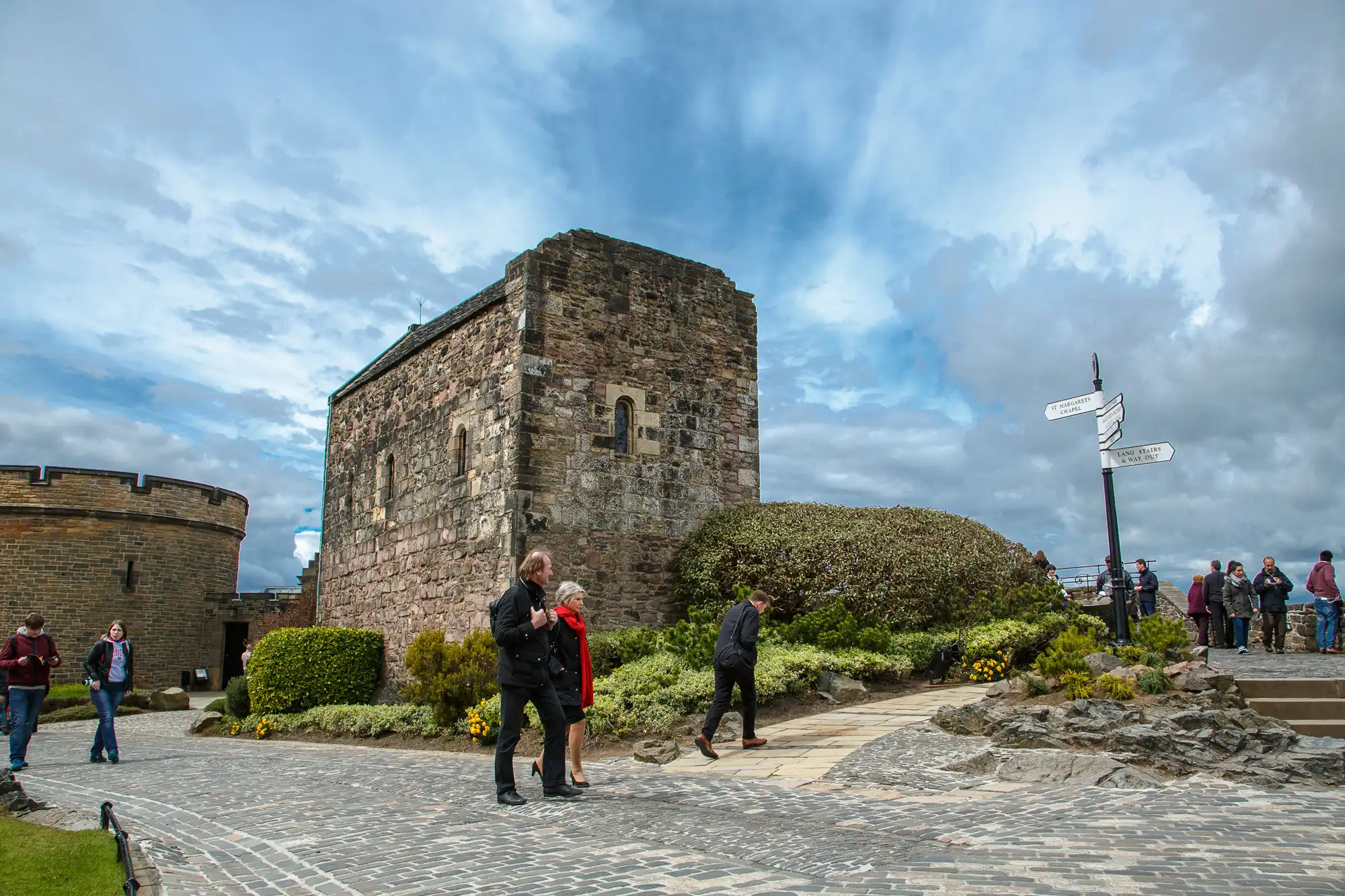 Visitors explore the grounds near an ancient stone building under a partly cloudy sky, with directional signposts and lush greenery visible.
