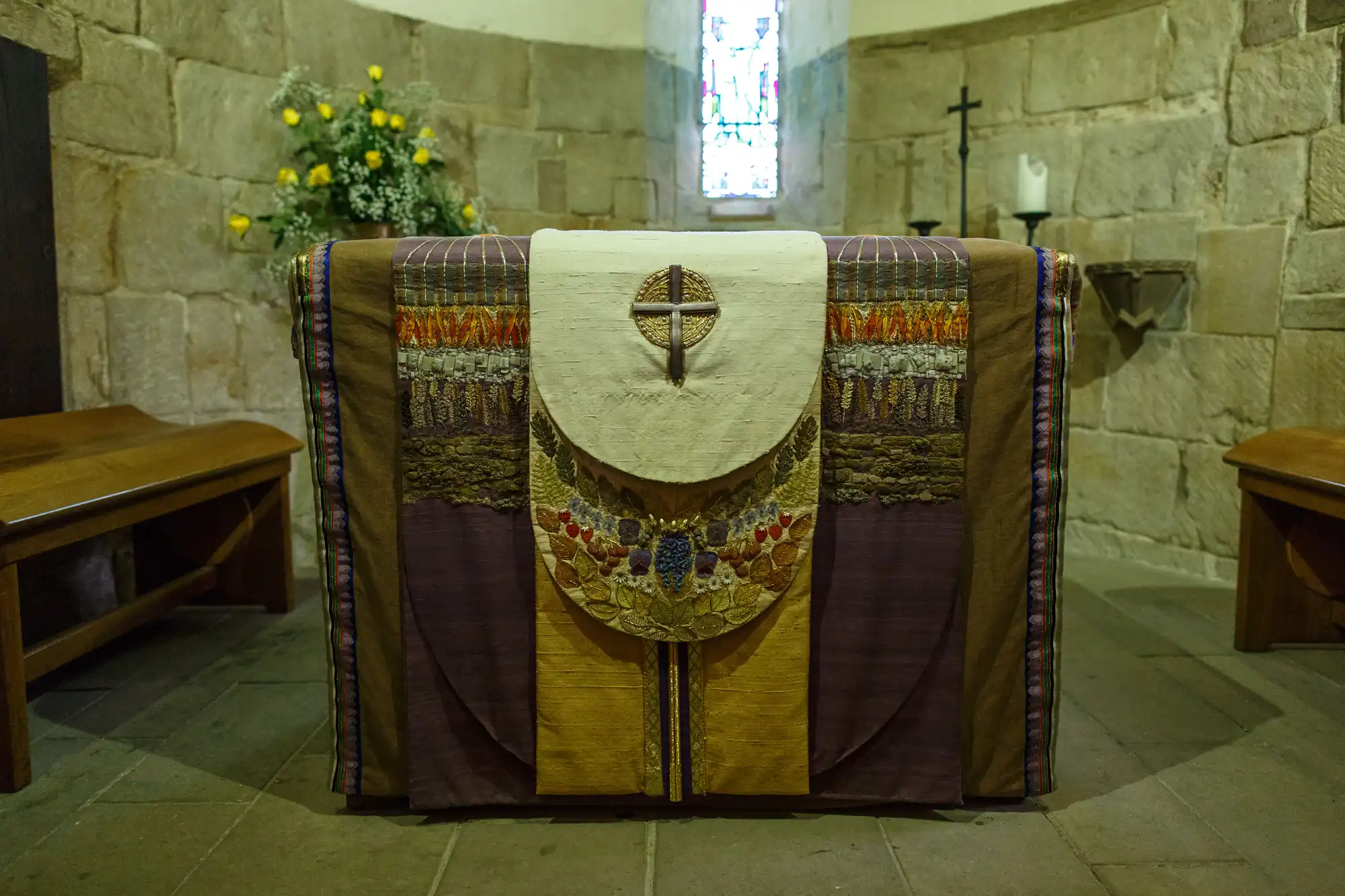 Altar with a decorative cloth and a cross emblem in the center, situated in a small, stone-walled chapel with a stained glass window in the background.