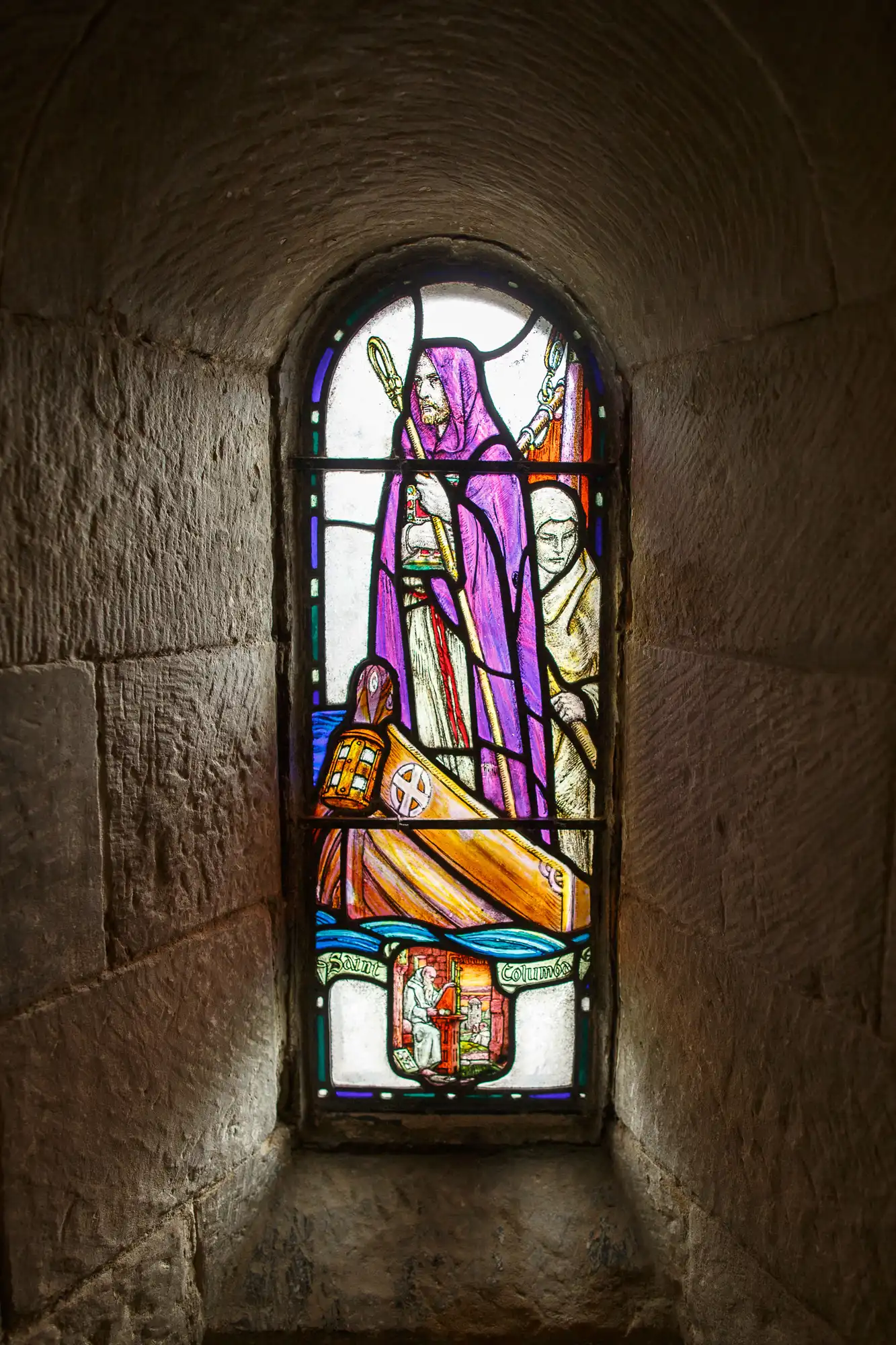 Stained glass window depicting a bishop in purple robes holding a staff, with another figure in the background, set in a stone arch.