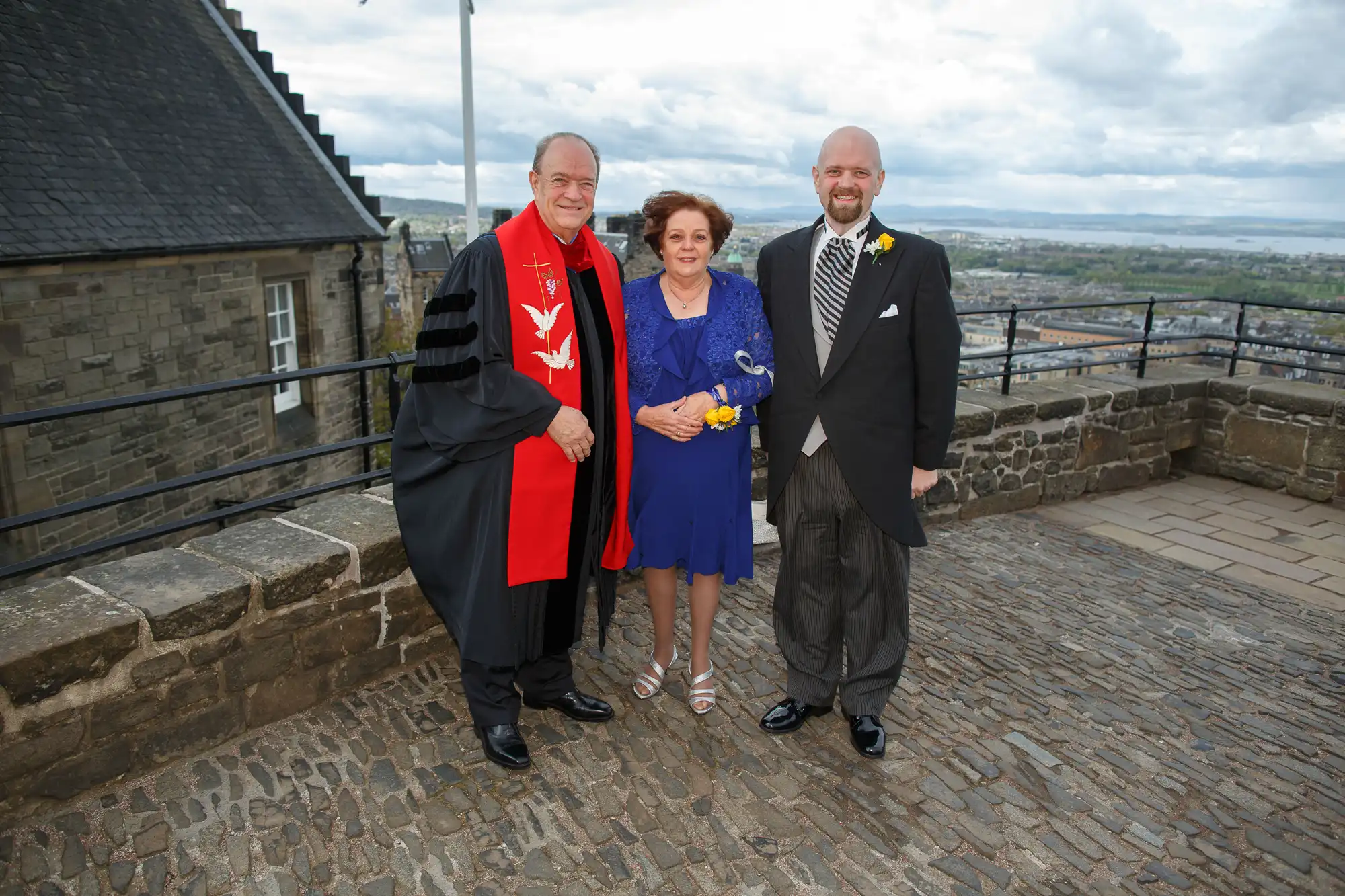 Three people, one in a red robe, stand together on a castle balcony overlooking a cityscape.