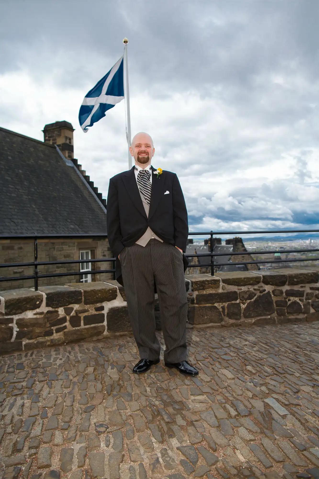 A man in a suit standing in front of a scottish flag at edinburgh castle with cloudy skies in the background.