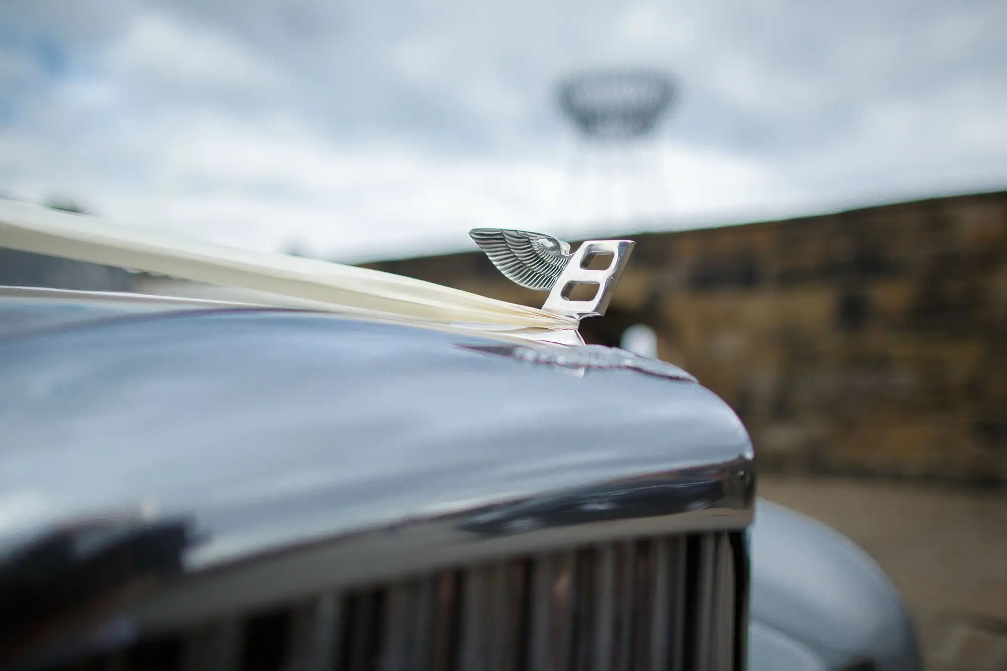 Close-up of a vintage car's hood ornament and grille, with a blurred background of a tower-like structure.
