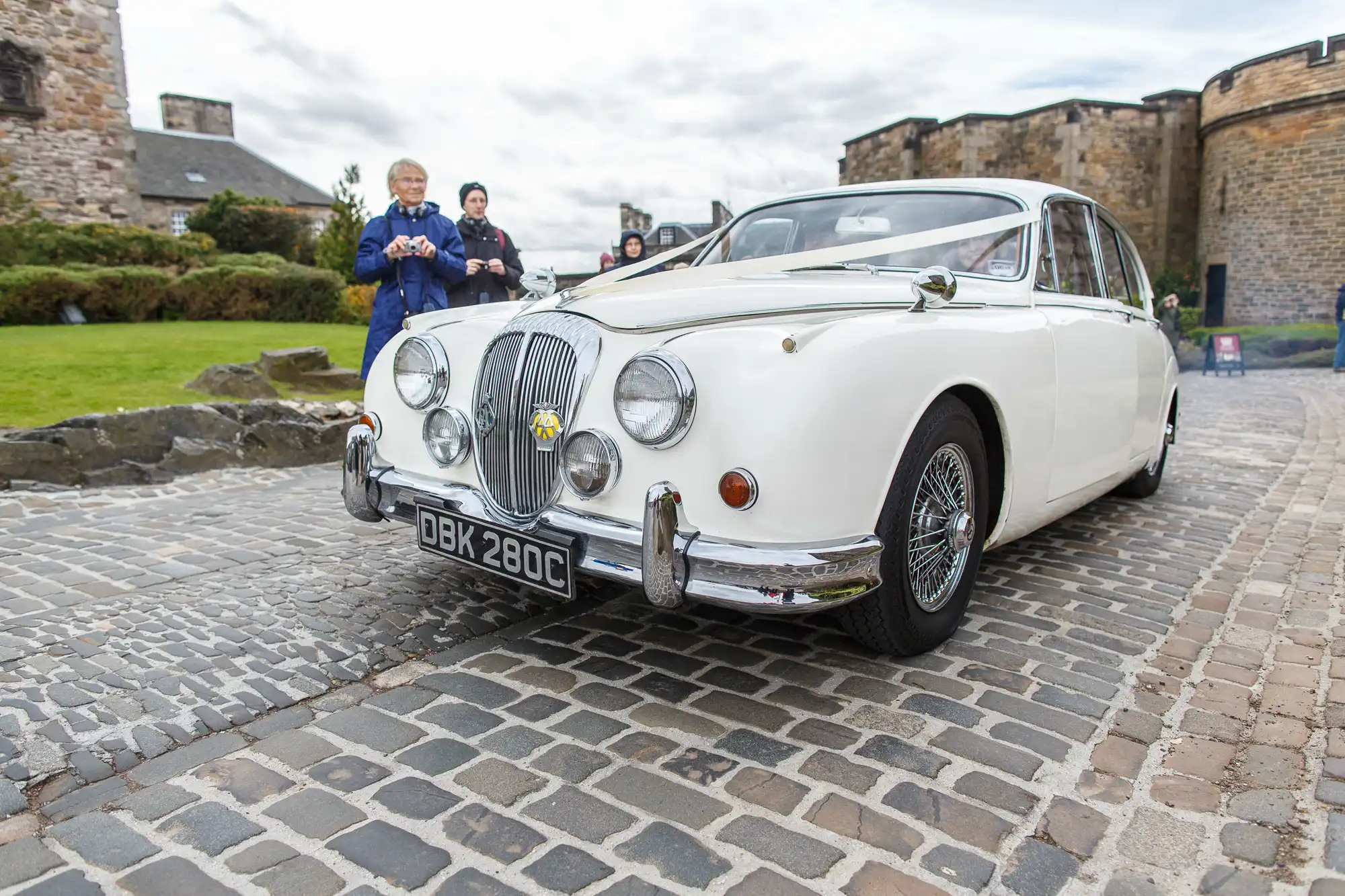 A vintage white jaguar car parked on cobblestones with a castle in the background and two people, one standing and one inside the car, dressed in formal wear.