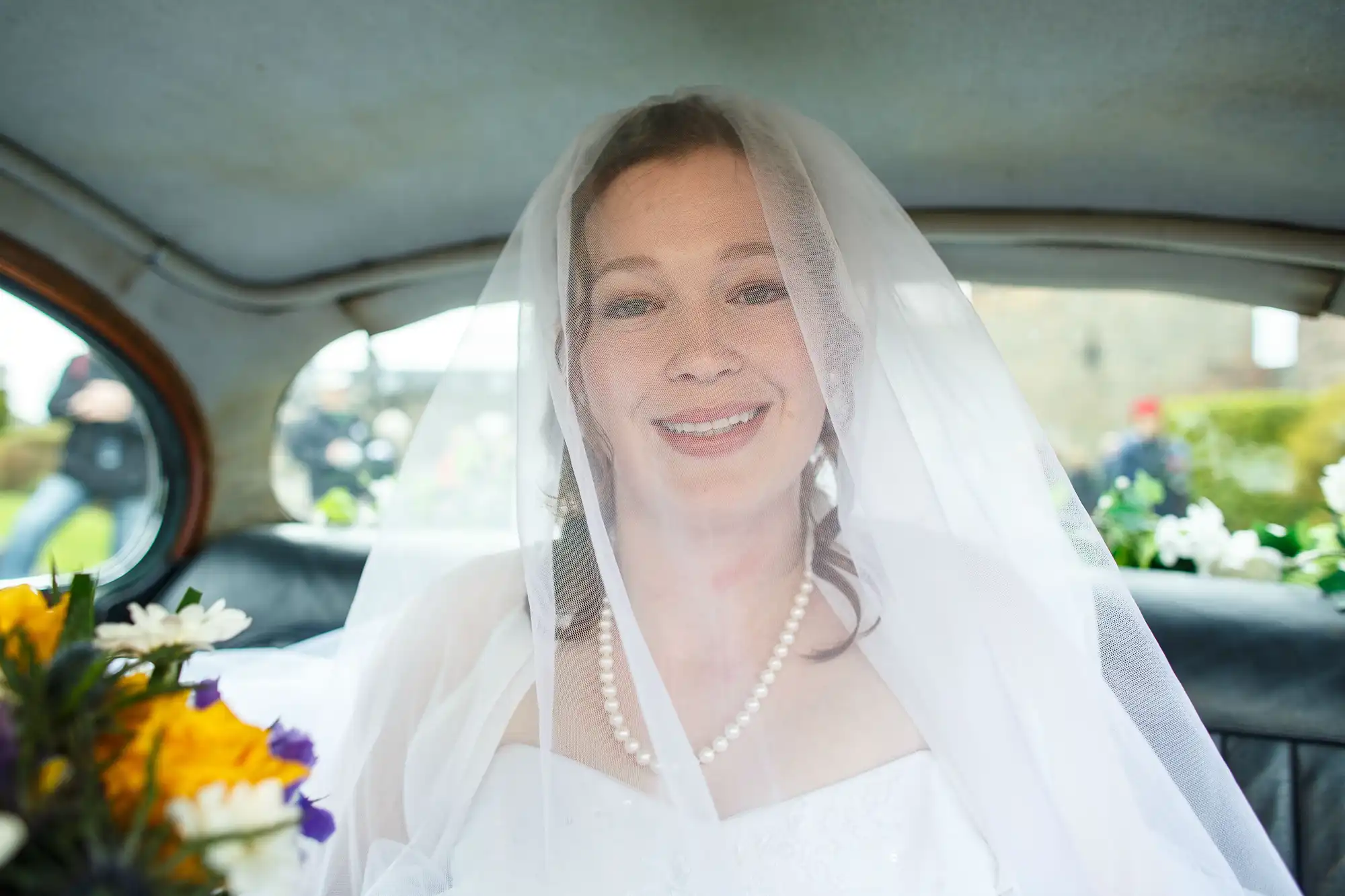 A bride in a wedding dress and veil smiles inside a vintage car, with visible flowers on the dashboard.