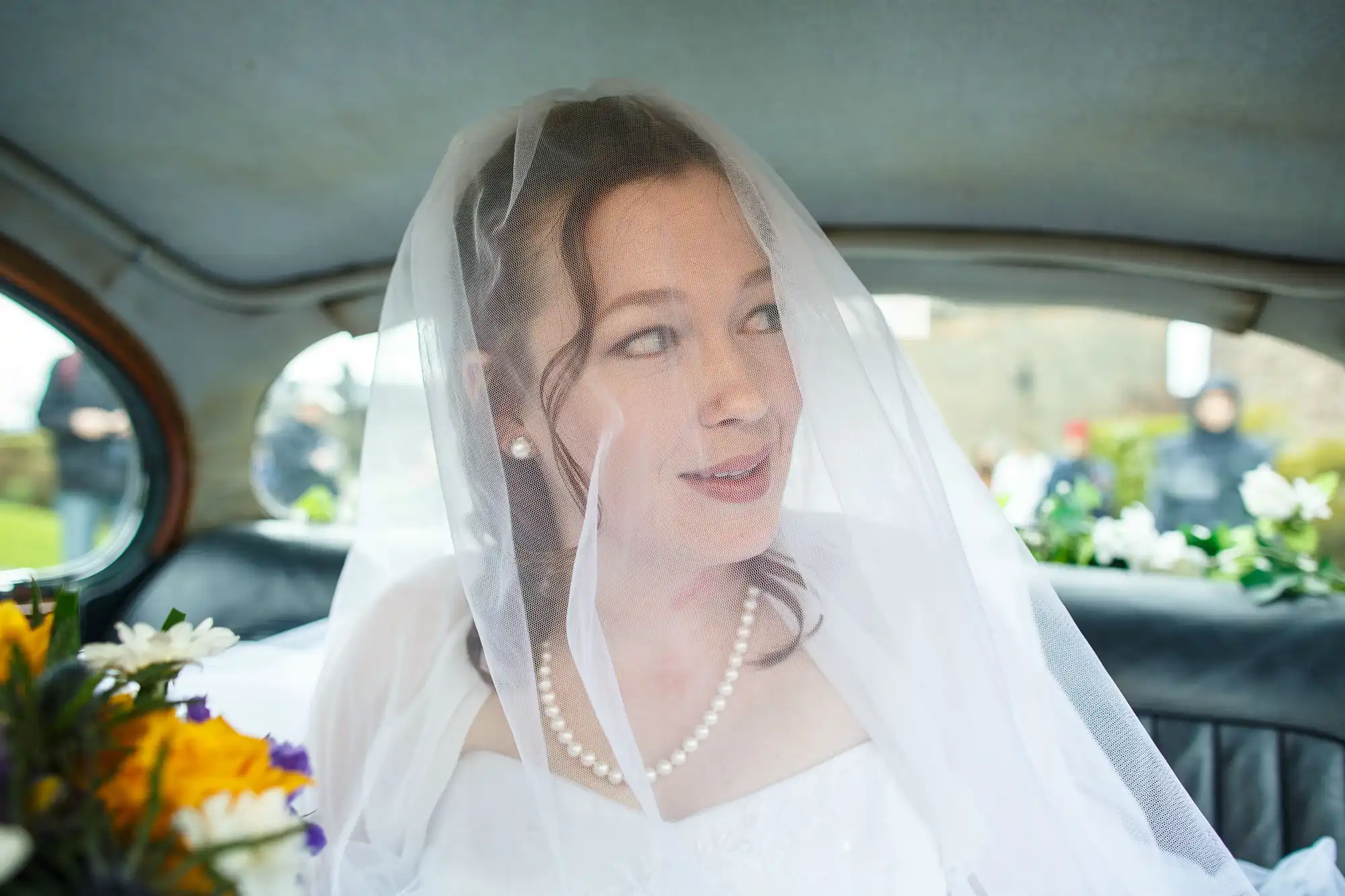 Bride in veil smiling inside a car, with flowers visible through the window.