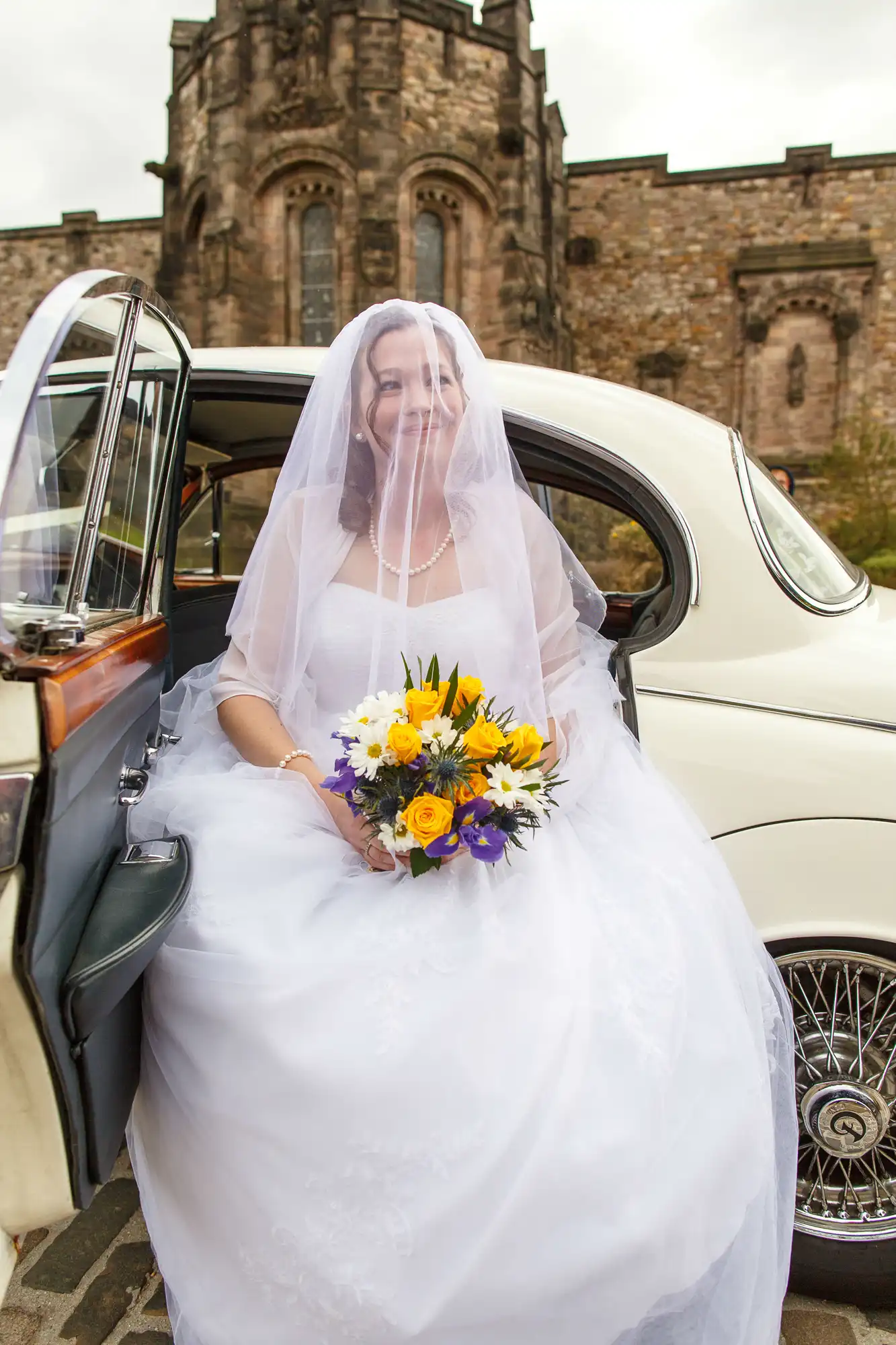 A bride in a white dress and veil sits in a classic car, holding a bouquet of yellow and purple flowers, with a historic castle in the background.