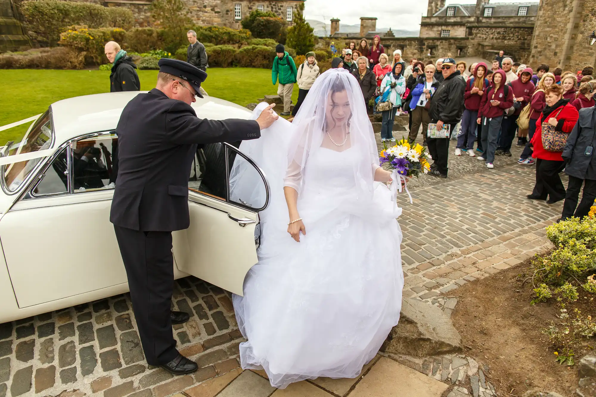 A bride in a white dress alighting from a classic car, assisted by a chauffeur in uniform, with onlookers in the background at an outdoor venue.