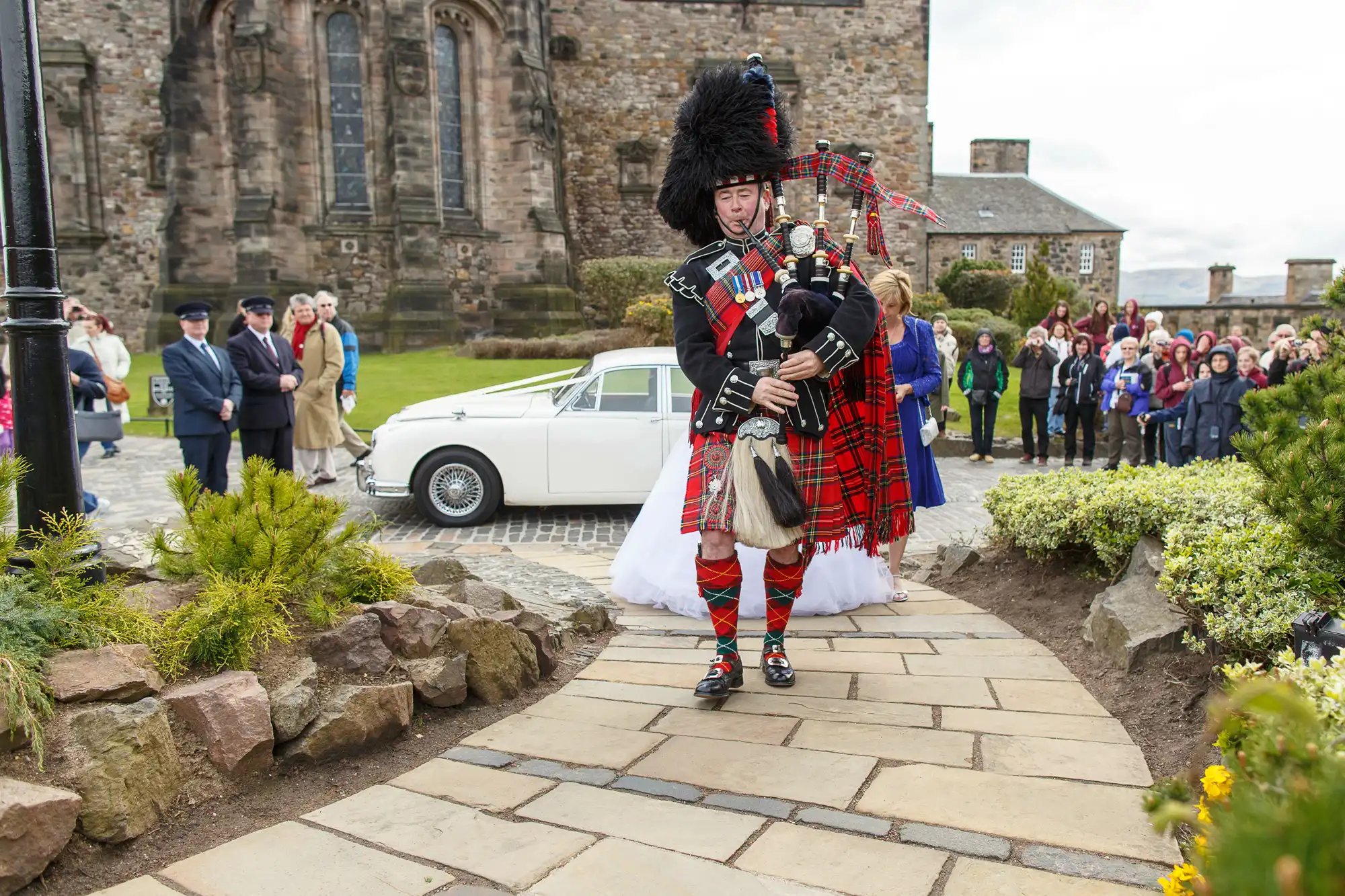 A scottish bagpiper in traditional attire leads a bride towards a vintage car, with onlookers gathered around in a historic setting.