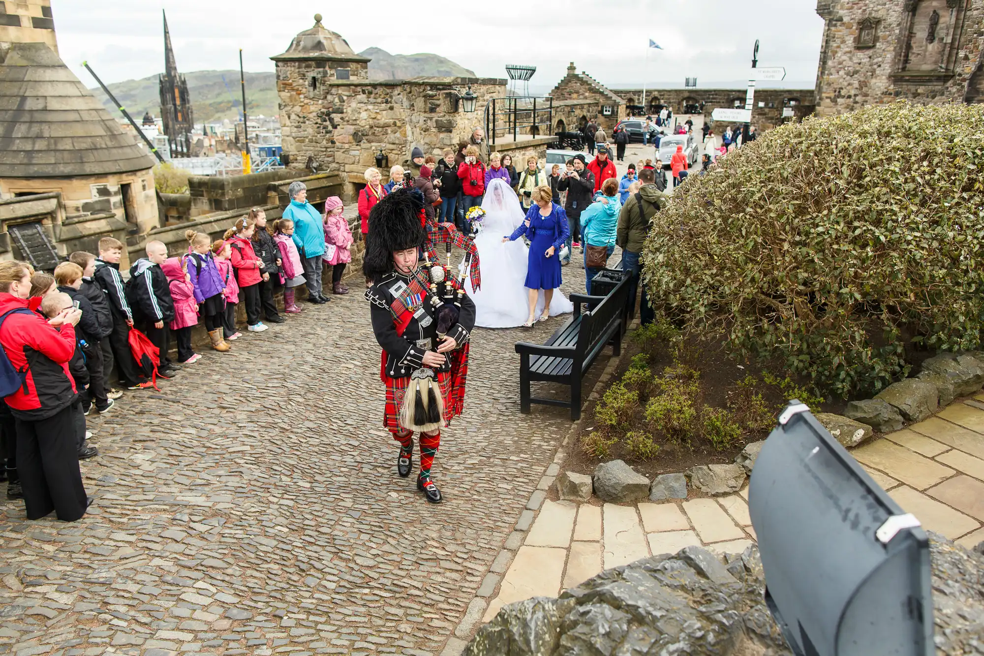 A bagpiper in traditional scottish attire walks past a crowd and a bride at edinburgh castle.