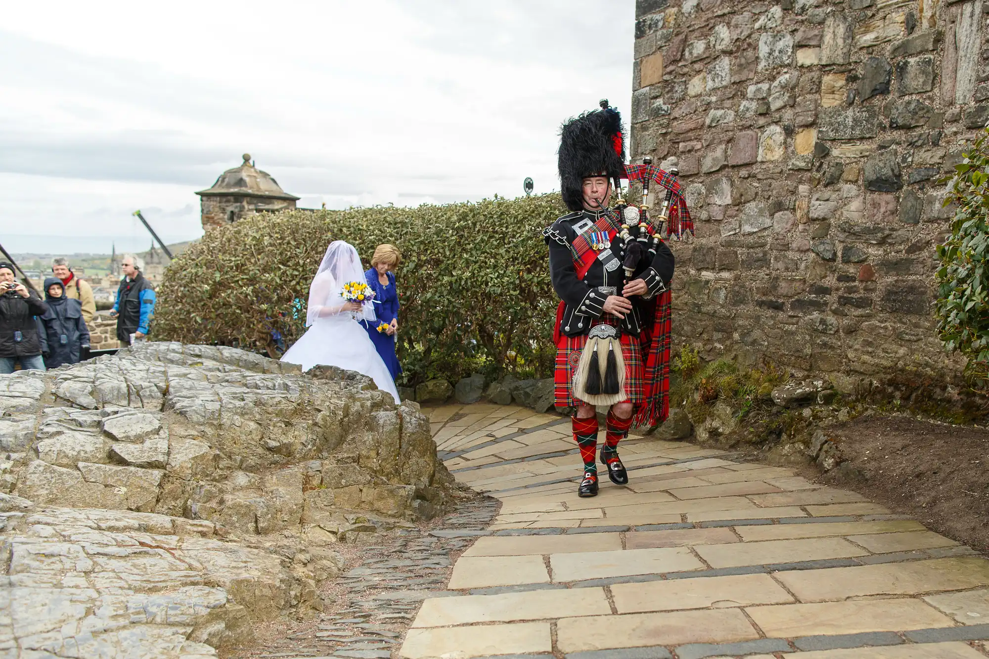 A bagpiper in traditional scottish attire leads a bride along a stone path at an outdoor wedding ceremony.