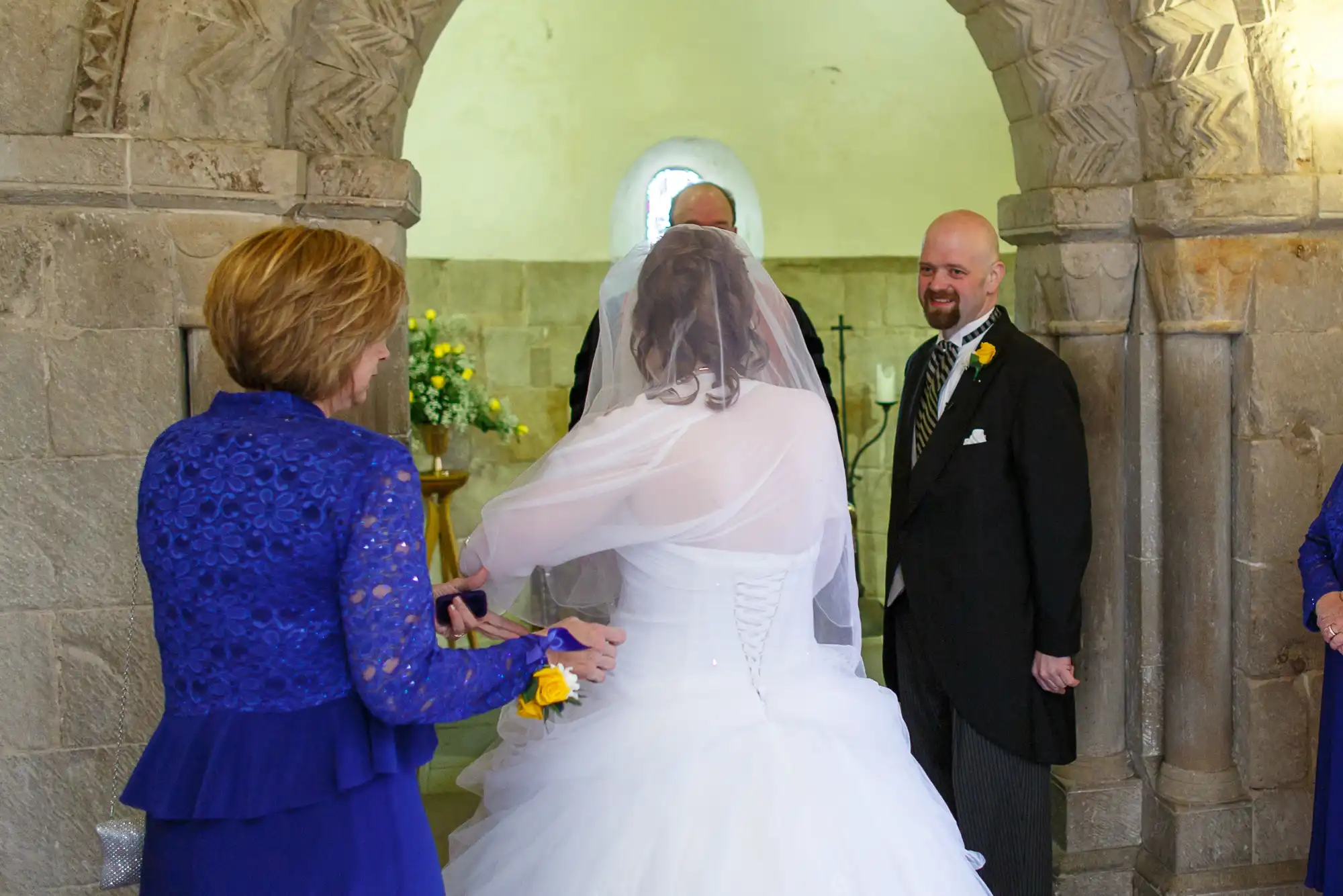 A bride in a white dress exchanges rings with her groom, who is smiling in a church ceremony.