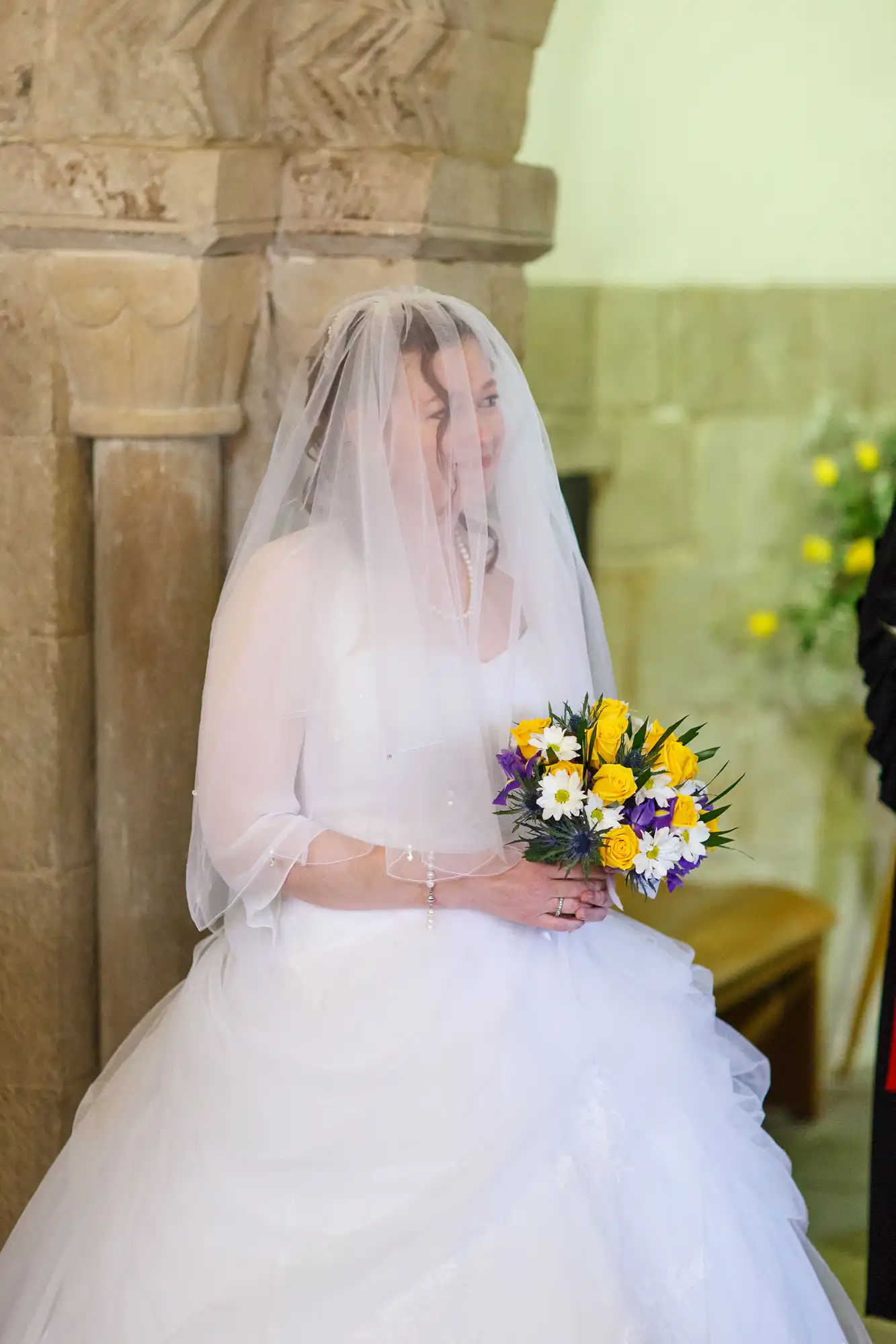 A bride in a white dress and veil holding a bouquet of yellow and purple flowers, smiling softly inside a church.