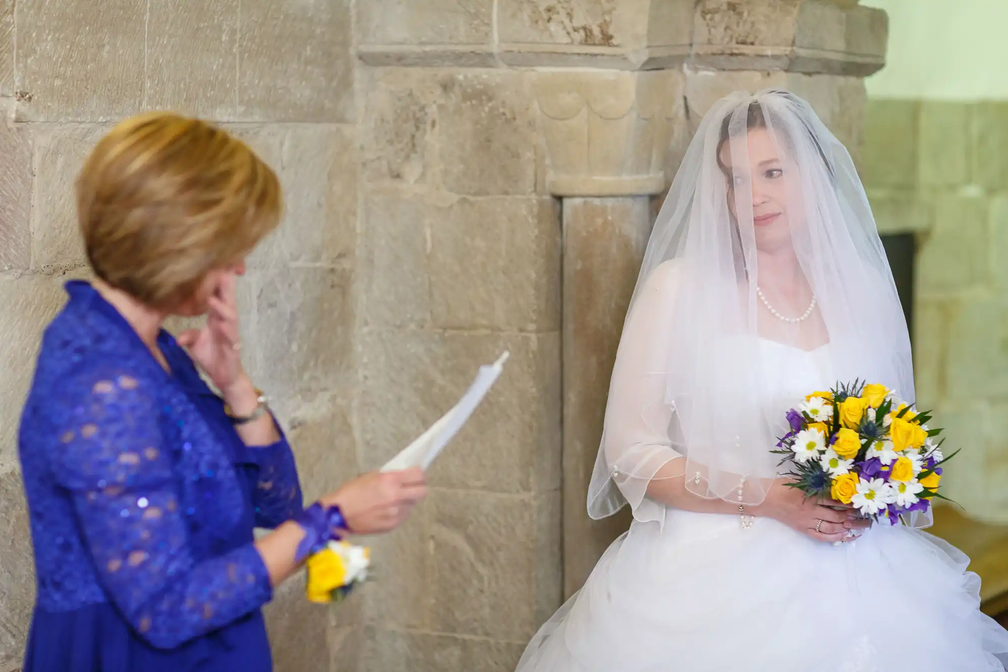 A bride in a white dress holds a bouquet while listening to an emotional woman in a blue dress reading from a paper in a stone church.