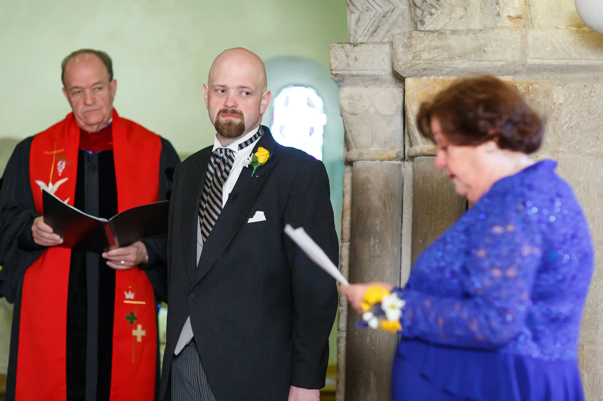 A groom stands with a priest and a woman, all holding papers in a church, during a wedding ceremony.
