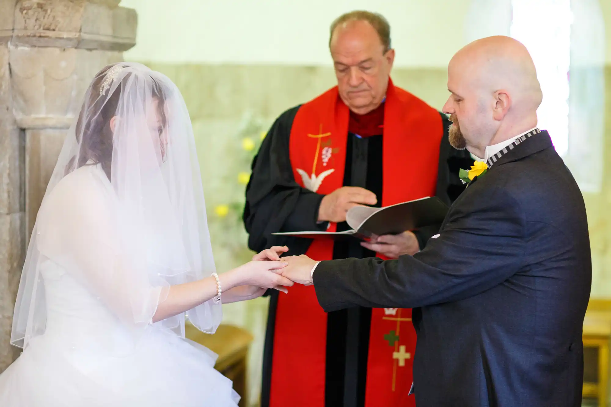 A bride and groom exchange rings during their wedding ceremony, officiated by a priest in a red robe, inside a church.