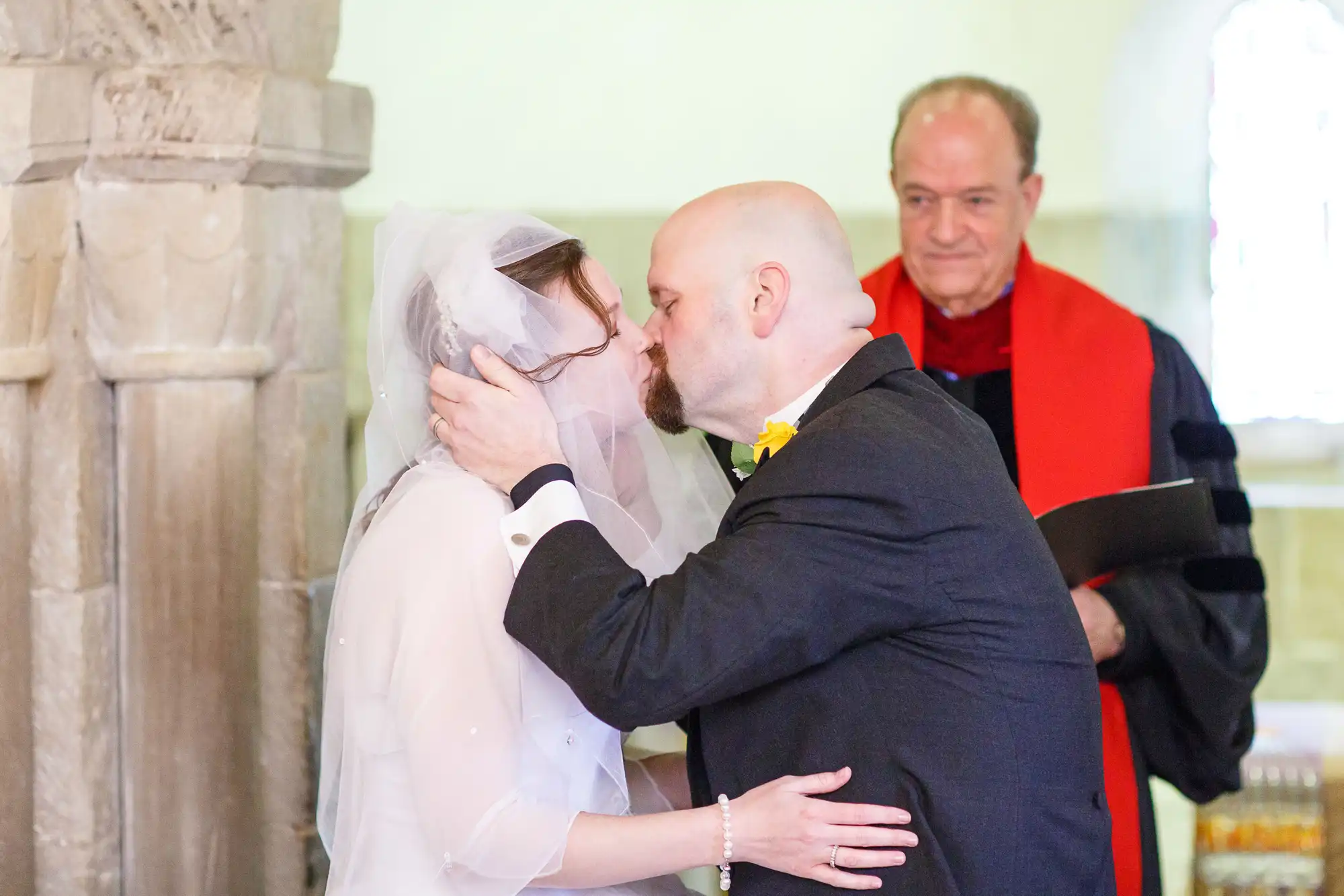 A bride and groom kiss at the altar, a priest in red robes observes in the background, in a church setting.