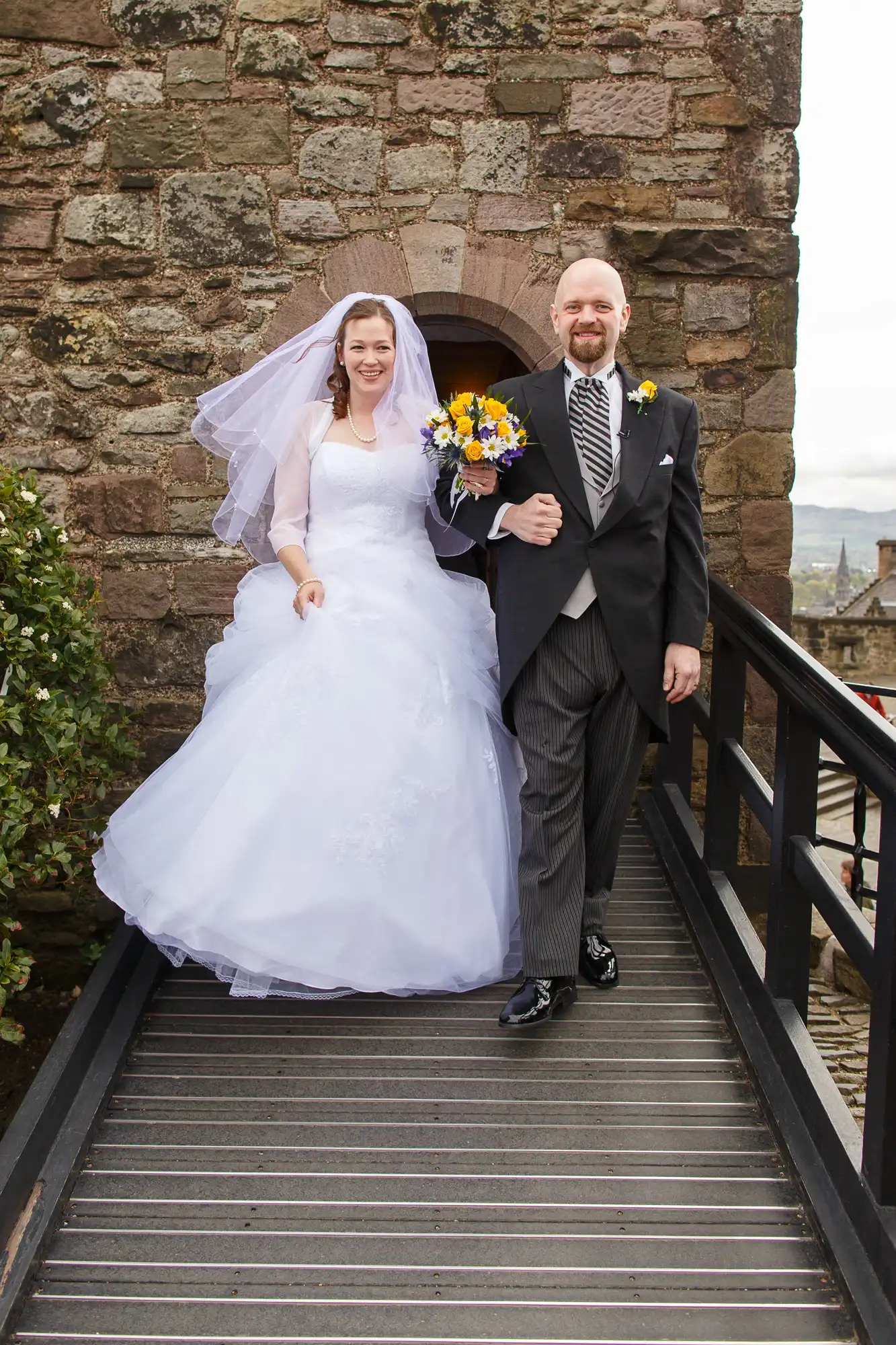 A smiling bride and groom walking down stairs, the bride in a white gown and veil and the groom in a gray suit, holding hands and a bouquet of colorful flowers.