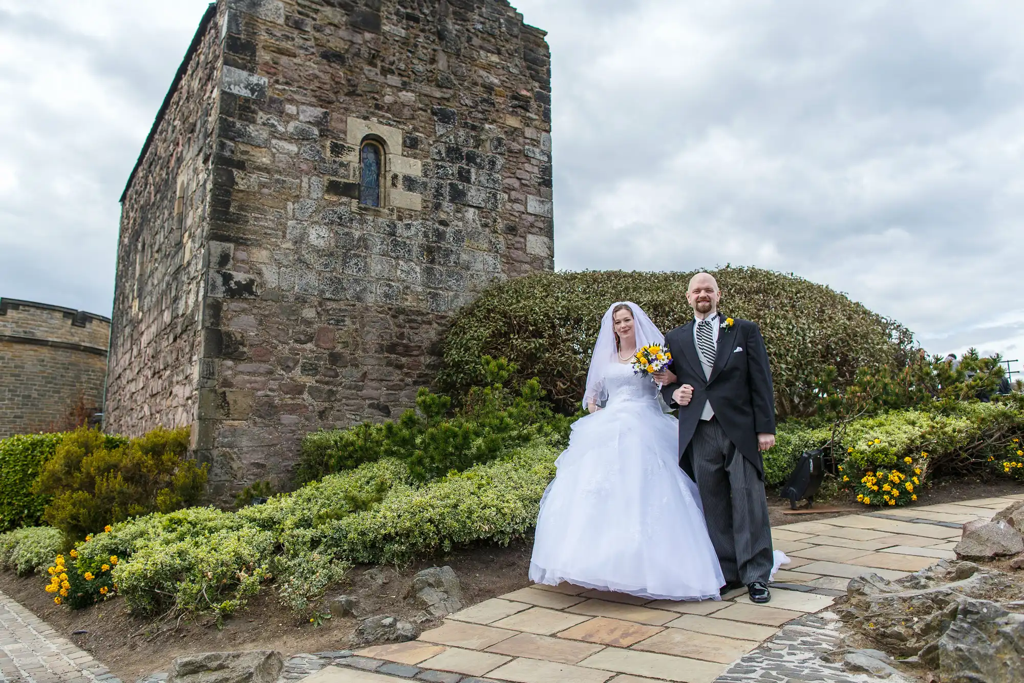 A bride in a white gown and a groom in a suit holding hands in a garden with a historic stone tower in the background.