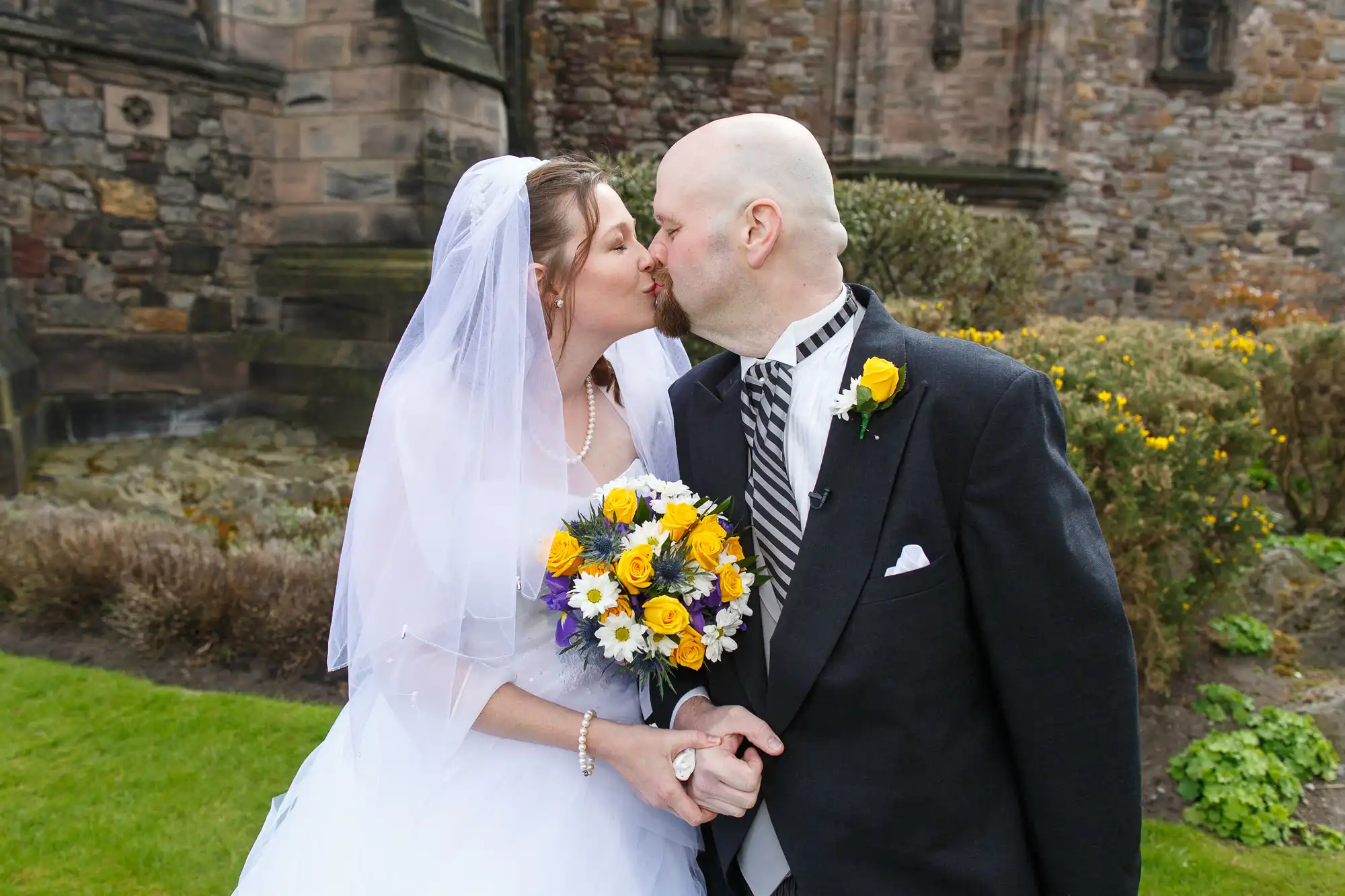 A bride and groom kiss outside a historic stone building, the bride holding a bouquet of yellow and blue flowers.