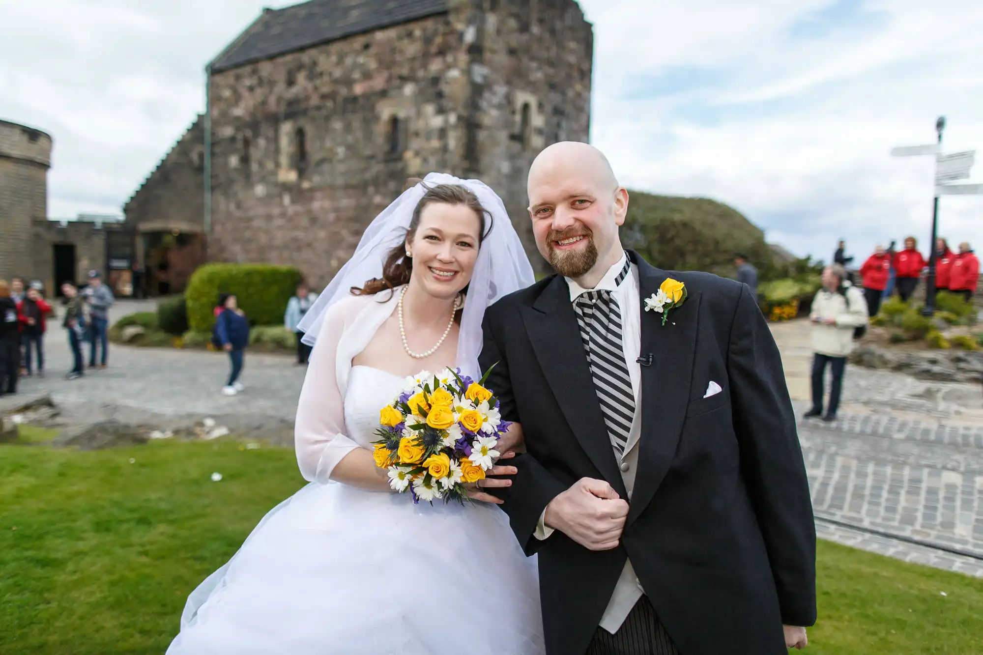 A bride and groom smiling, holding hands in front of an old stone church, the bride holding a bouquet of yellow flowers.