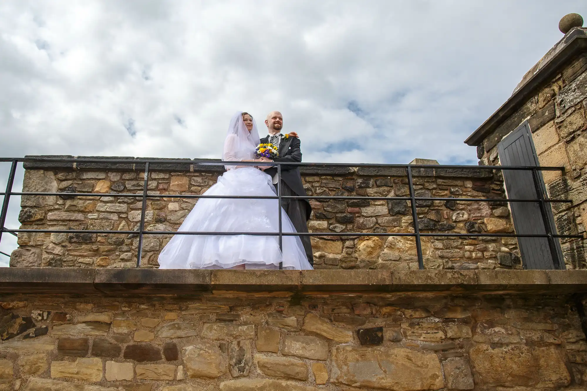 A bride in a white dress and a groom in a suit holding a bouquet stand together on a stone wall under a cloudy sky.
