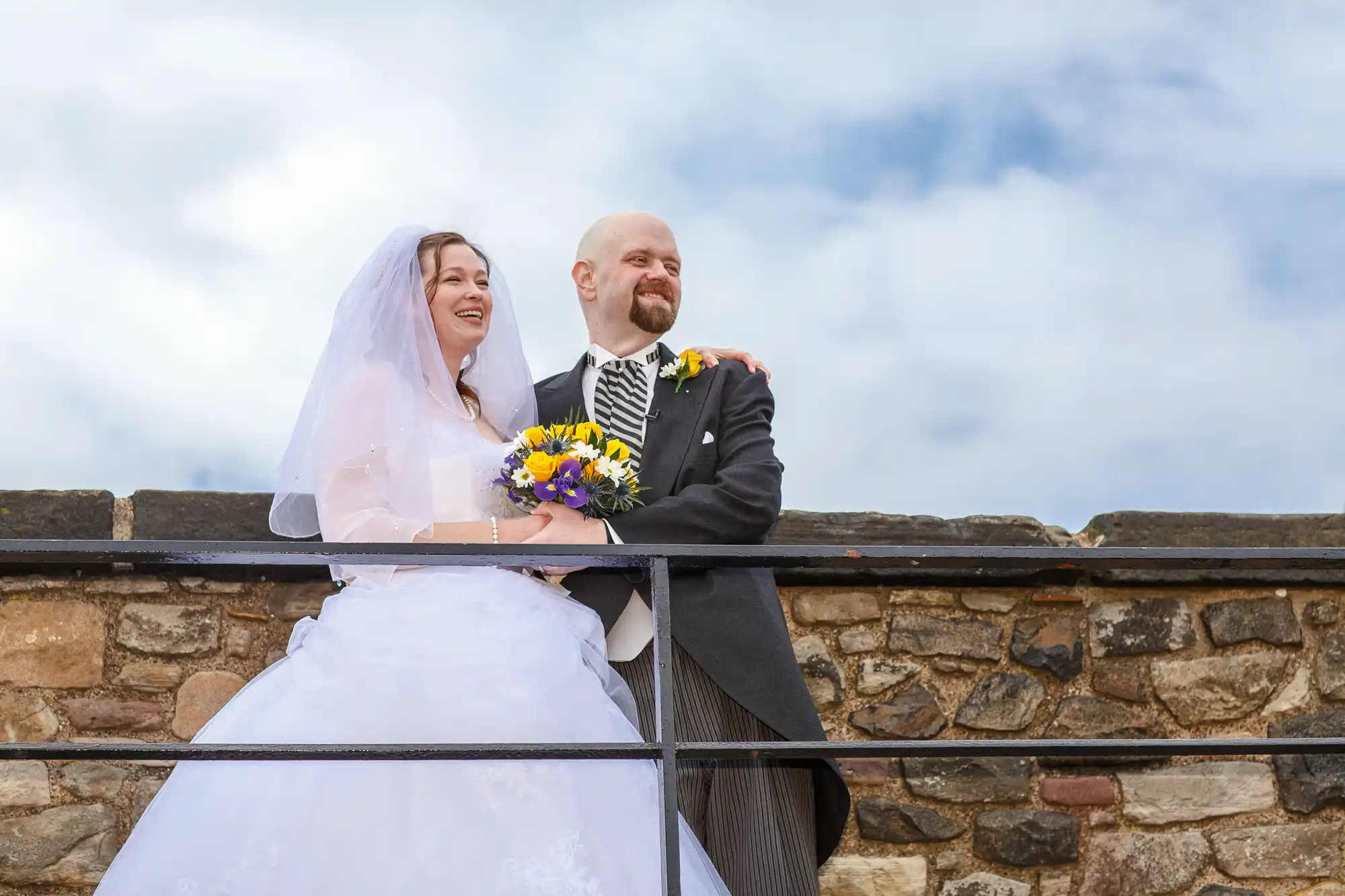 A joyous bride and groom, dressed in traditional wedding attire, laugh together on a stone balcony under a cloudy sky.