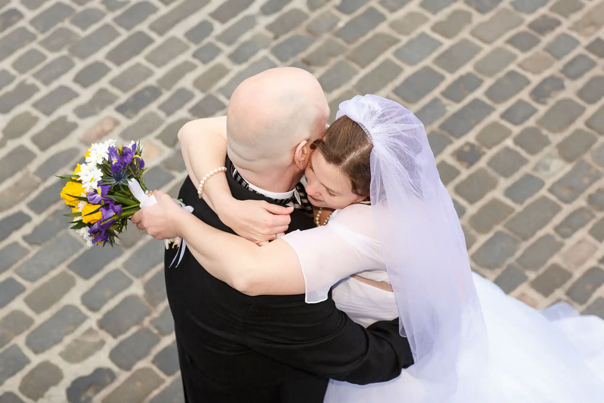 Bald groom in a black vest hugs a bride in a white dress and veil, holding a colorful bouquet, top-down view on a cobblestone street.