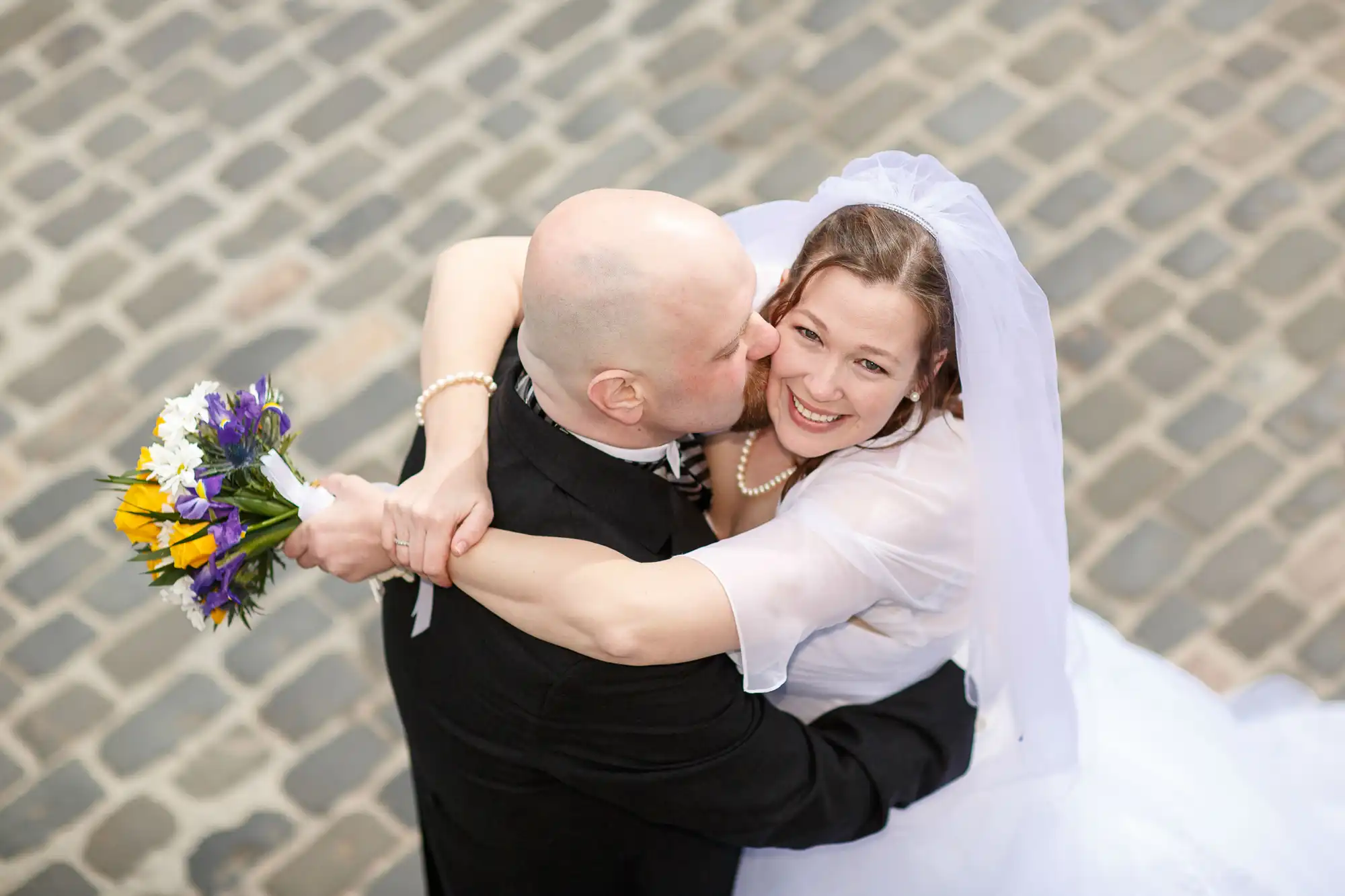 Bald groom in black suit kissing a smiling bride in a white dress with a veil, holding a bouquet, from an overhead perspective on a cobblestone street.