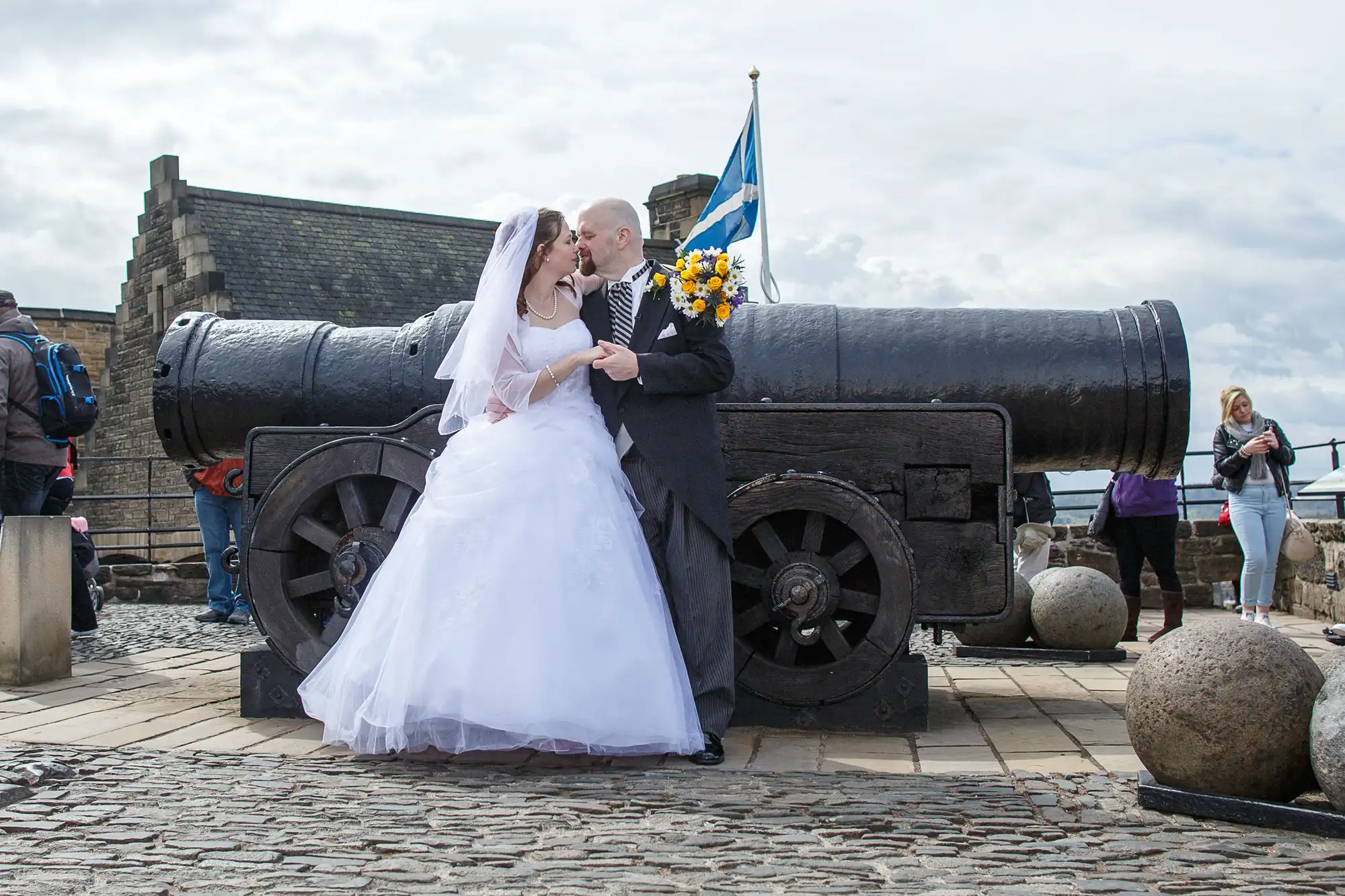 A bride and groom share a kiss beside a large cannon, with sunflowers and a scottish flag, at a historical stone fortress.