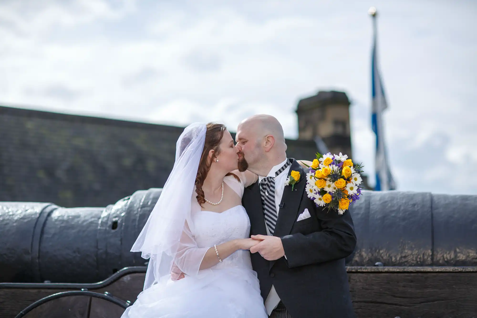 Bride and groom kissing beside a cannon, with a castle and a flag in the background, under a cloudy sky.