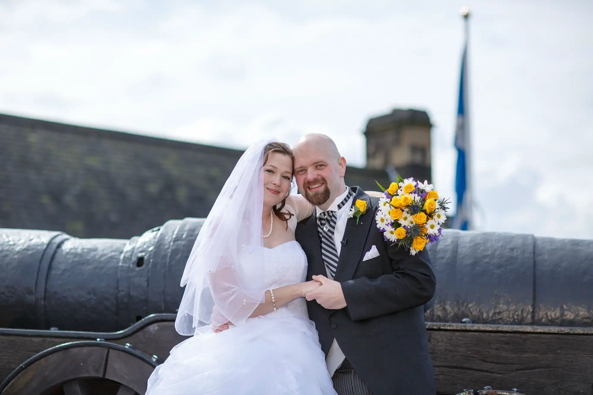 A bride and groom smiling at the camera, standing by an old cannon with a castle and a blue flag in the background. the bride holds a bouquet of yellow flowers.