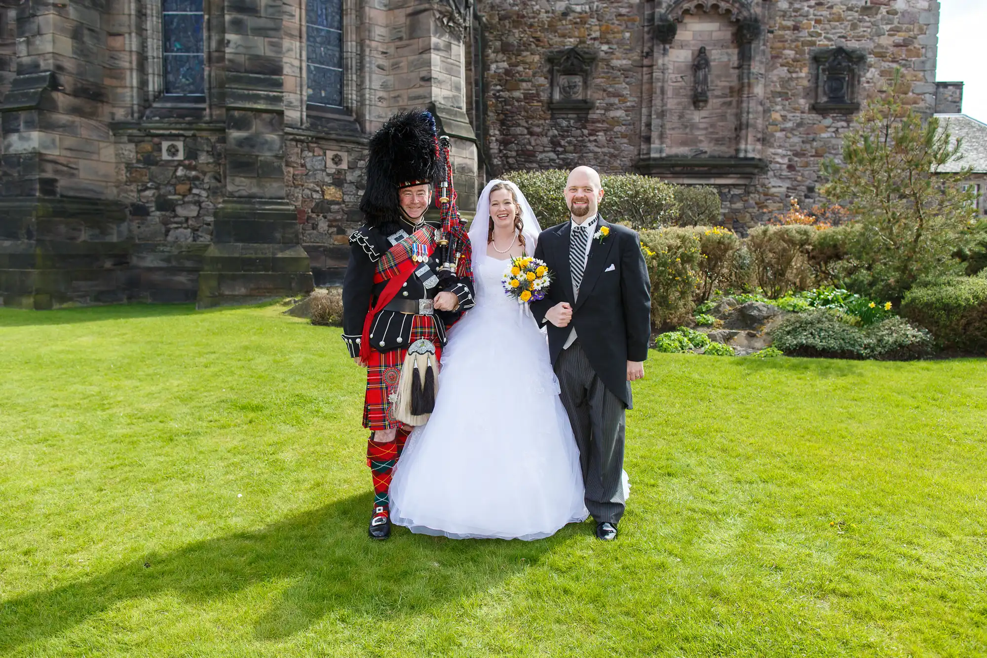 A bride and groom smiling with a bagpiper in traditional scottish attire, standing on a grassy field beside a historic stone building.