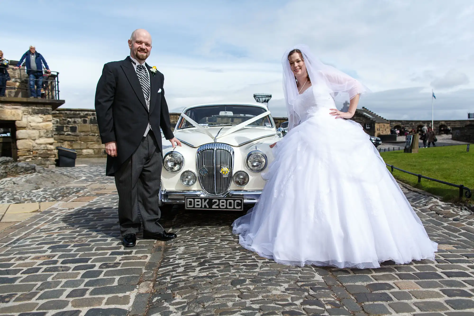 Bride and groom smiling beside a classic car on a stone pavement, with a historic castle and clear skies in the background.