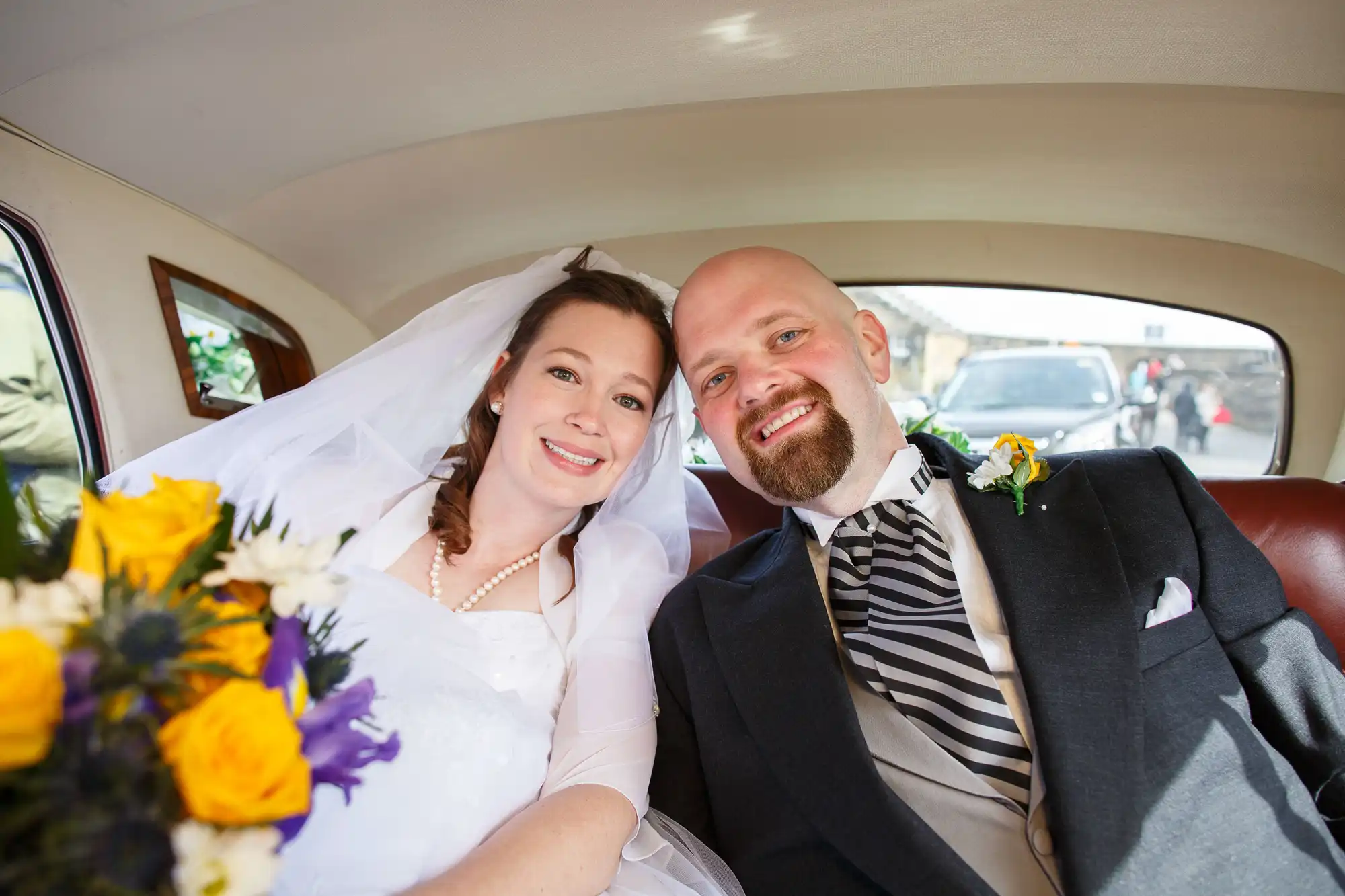 A newlywed couple smiling inside a vintage car, the bride in a white dress and veil, the groom in a striped suit, with a bouquet in the foreground.