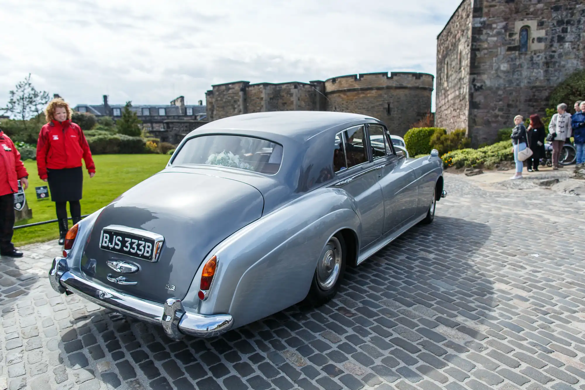 A vintage silver rolls-royce parked on cobblestones near a historic castle, with people walking around.