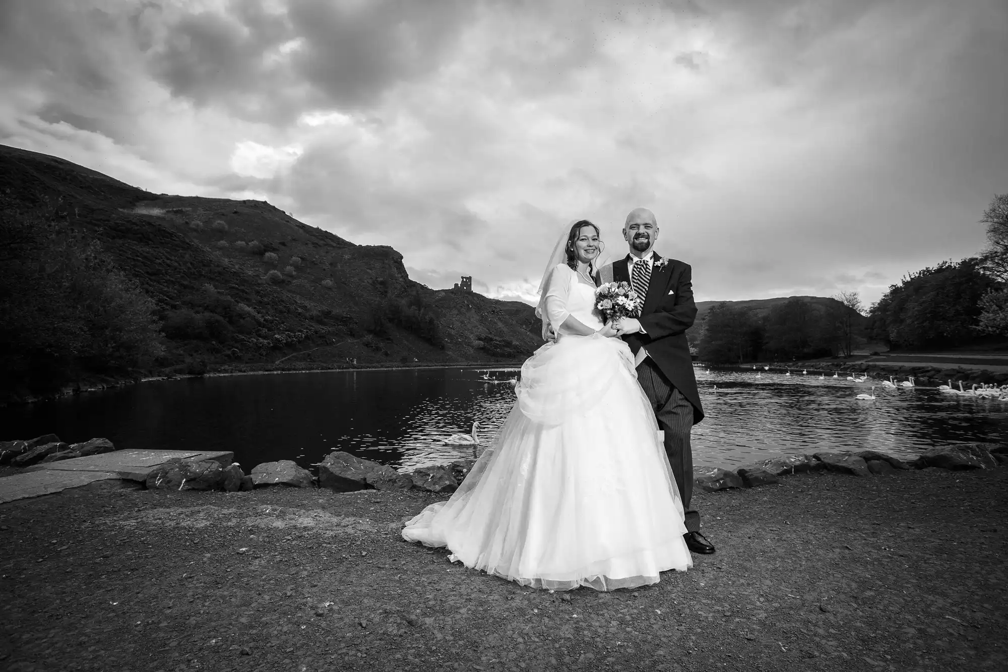 A bride and groom smiling along a lakeside, with rolling hills in the background under a cloudy sky, in a black and white photo.
