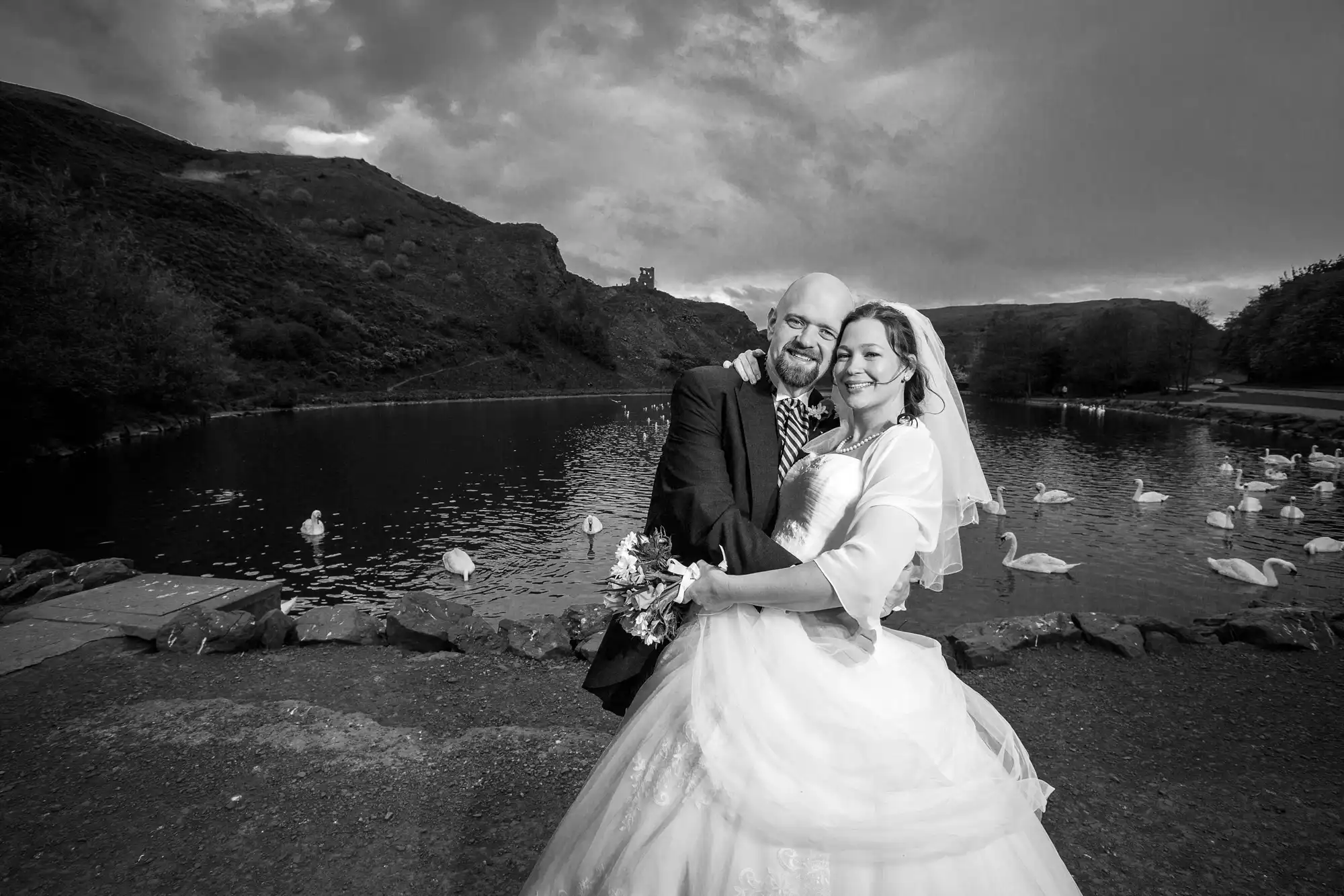 A bride and groom smiling by a lake with swans, under a dramatic sky and a distant castle on a hill.
