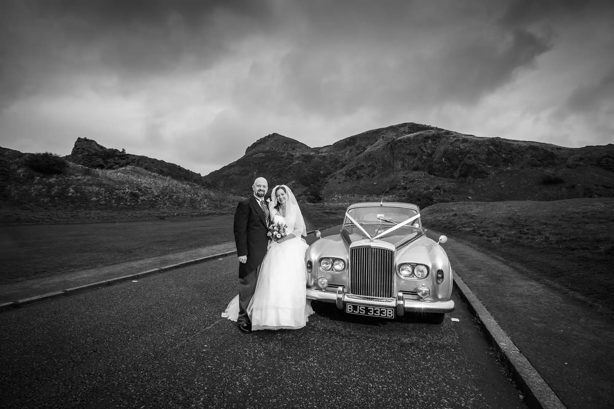 A bride and groom stand beside a classic car on a cloudy day in a mountainous landscape, both smiling at the camera.