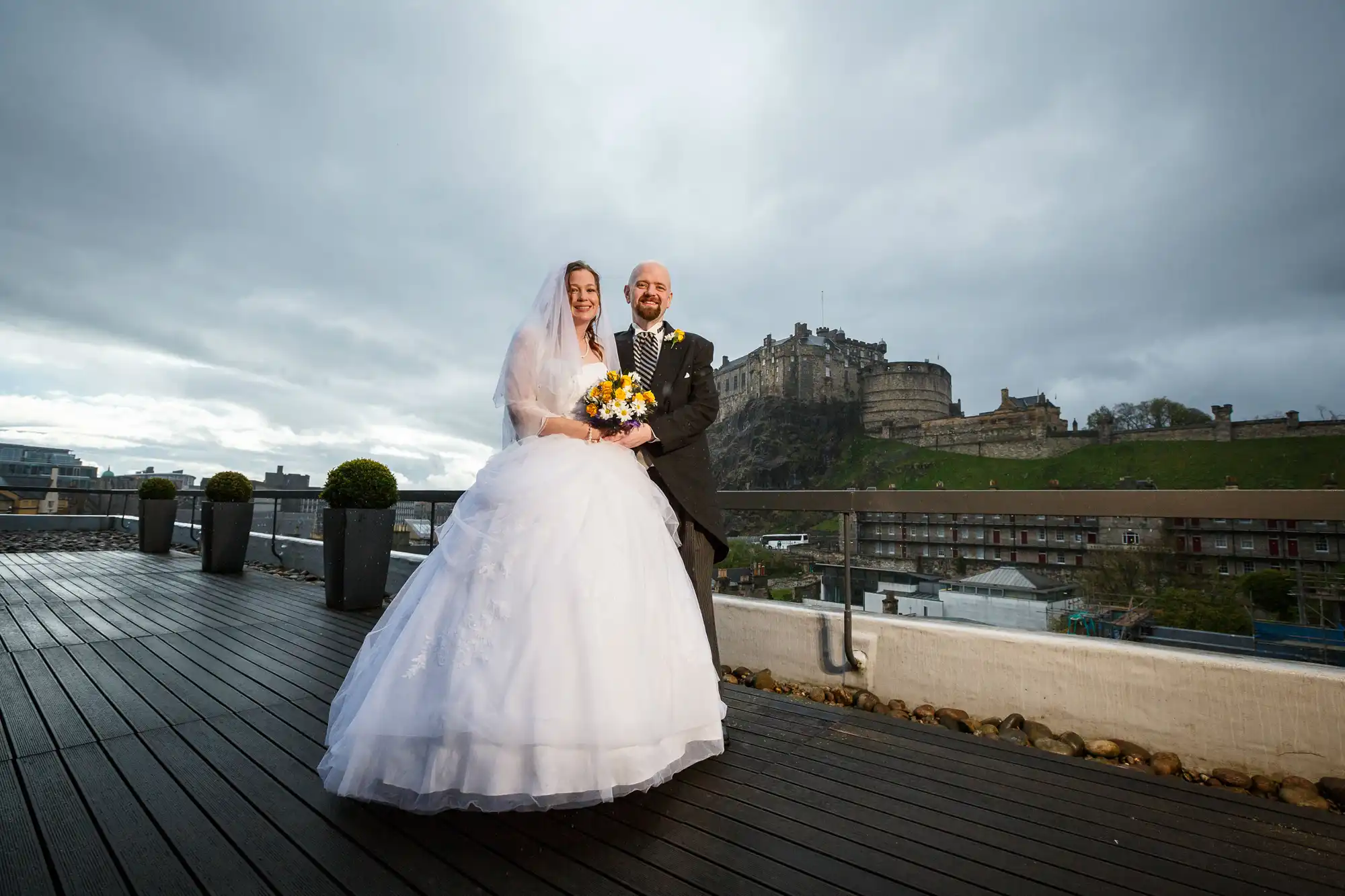 Bride and groom smiling on a rooftop, with a scenic view of edinburgh castle in the background, under a cloudy sky.