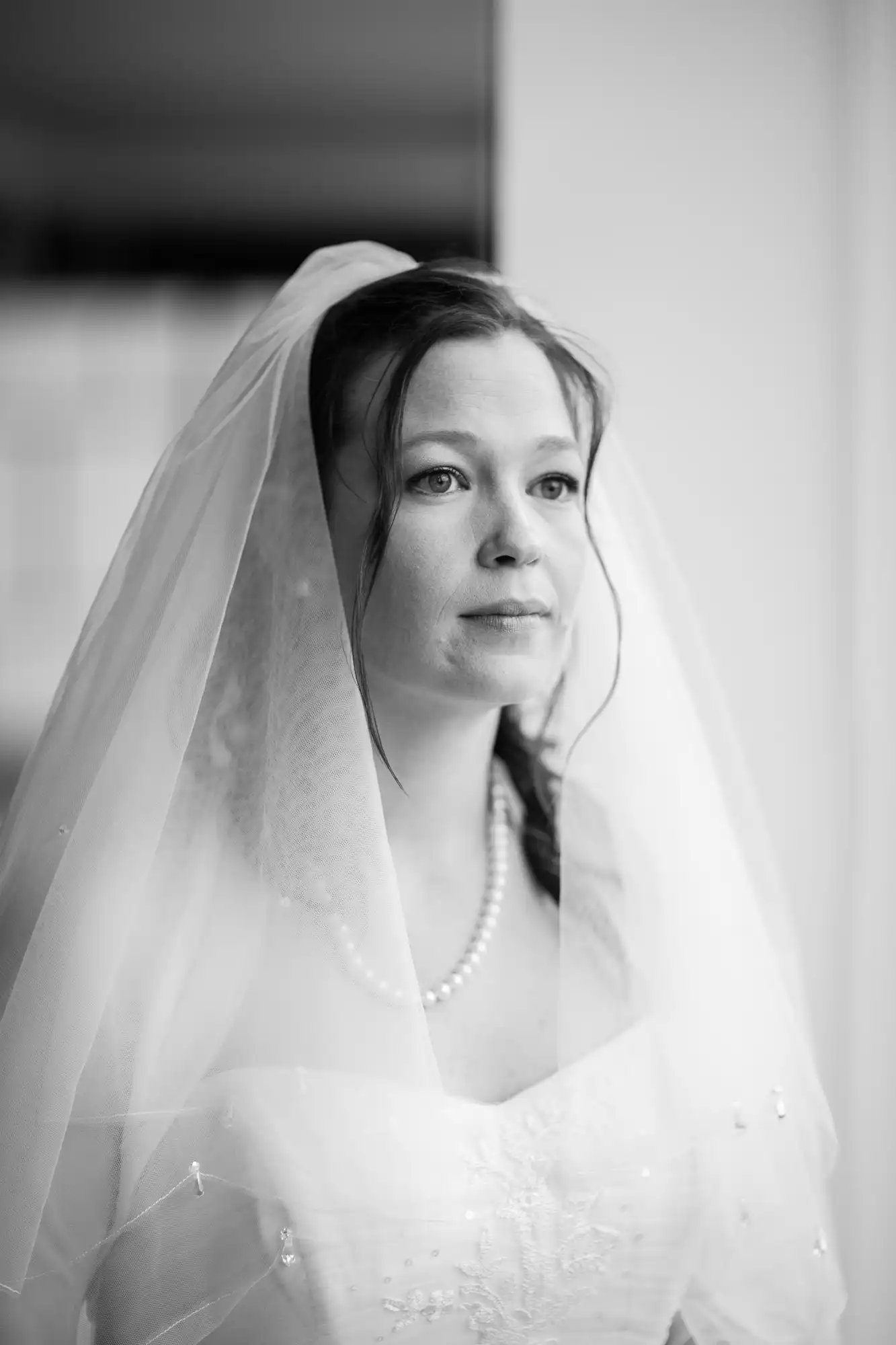 A bride in a white gown and veil, with pearls, looks pensively out a window in a softly lit black and white photograph.